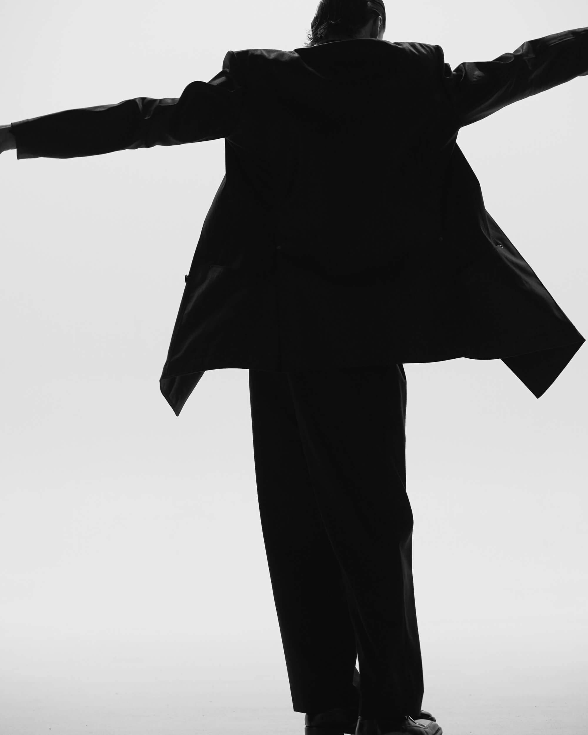 A person in a suit stands with arms extended sideways, seen from behind in a black-and-white photo with a plain, light background. The jacket is open and the figure is slightly leaning back.