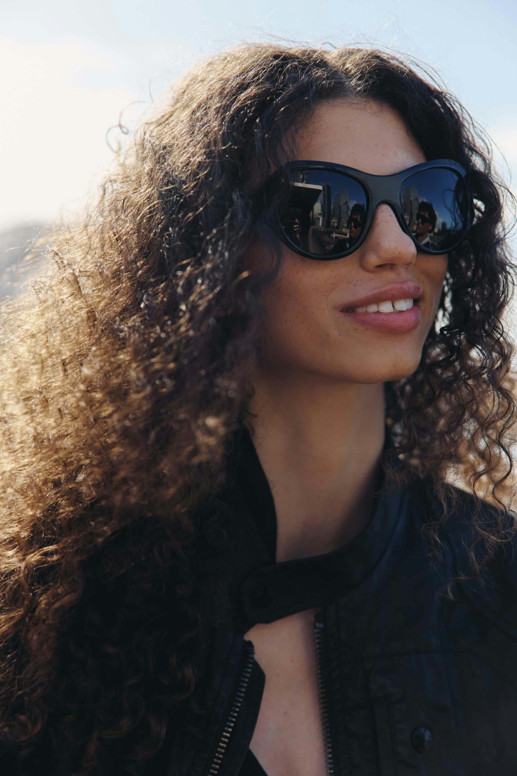 A person with long, curly hair wearing a black jacket and large, dark sunglasses is outdoors on a sunny day. The reflection of a cityscape is visible in the sunglasses.