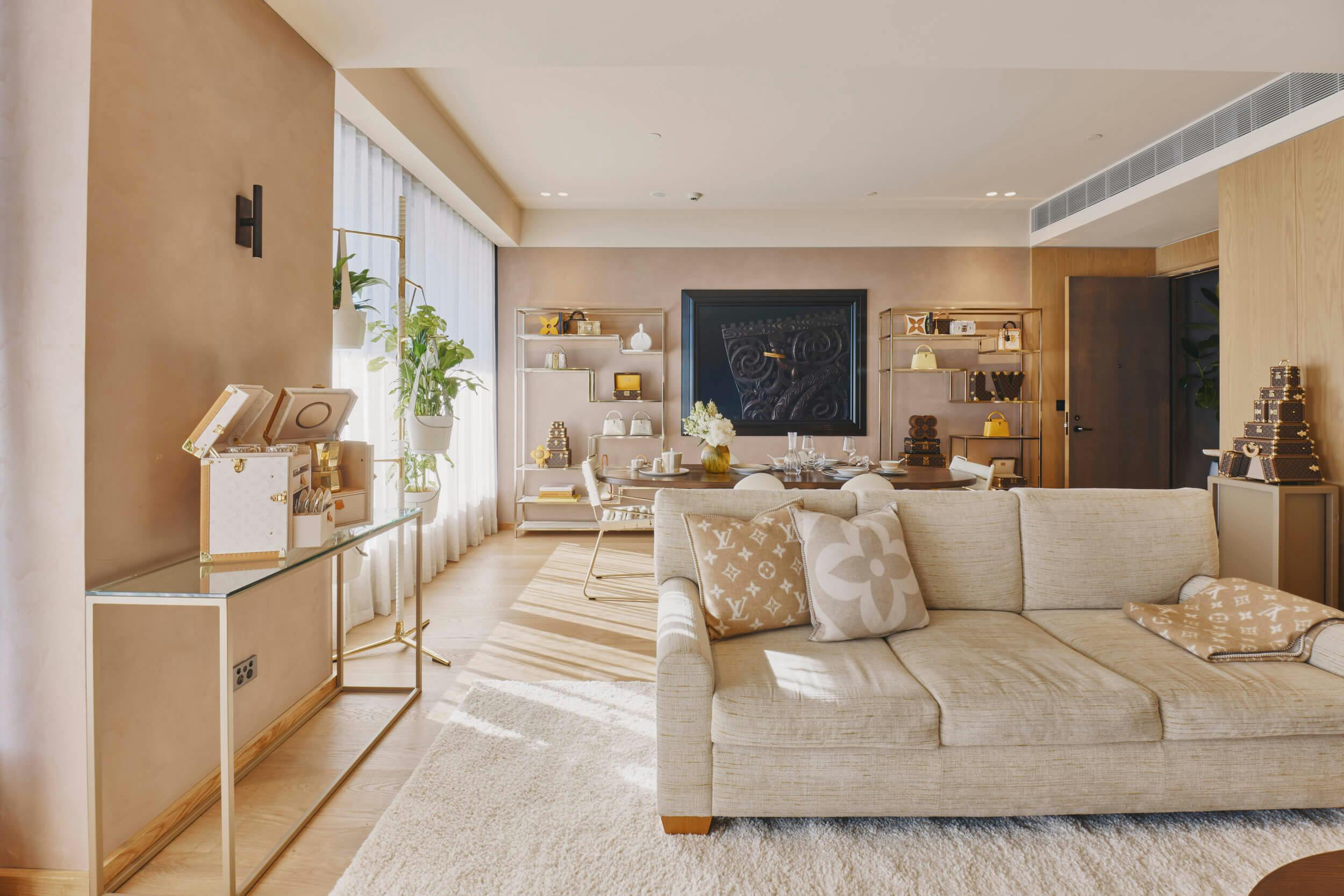 A modern living room with neutral tones features a beige sofa with patterned cushions, a glass console table, shelves with decor items, a dining area in the background, large windows, and indoor plants.