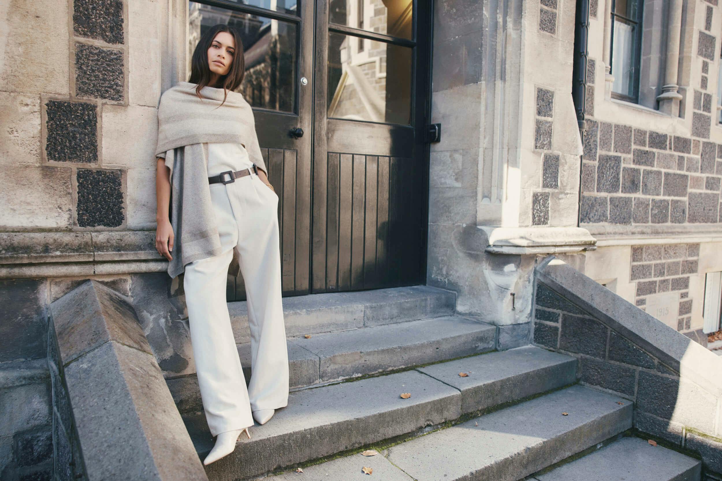 A woman stands on stone steps outside a building with a black door and stone walls. She is wearing a beige shawl, white wide-leg trousers, a belt, and white shoes. She is leaning against the wall with one hand in her pocket.