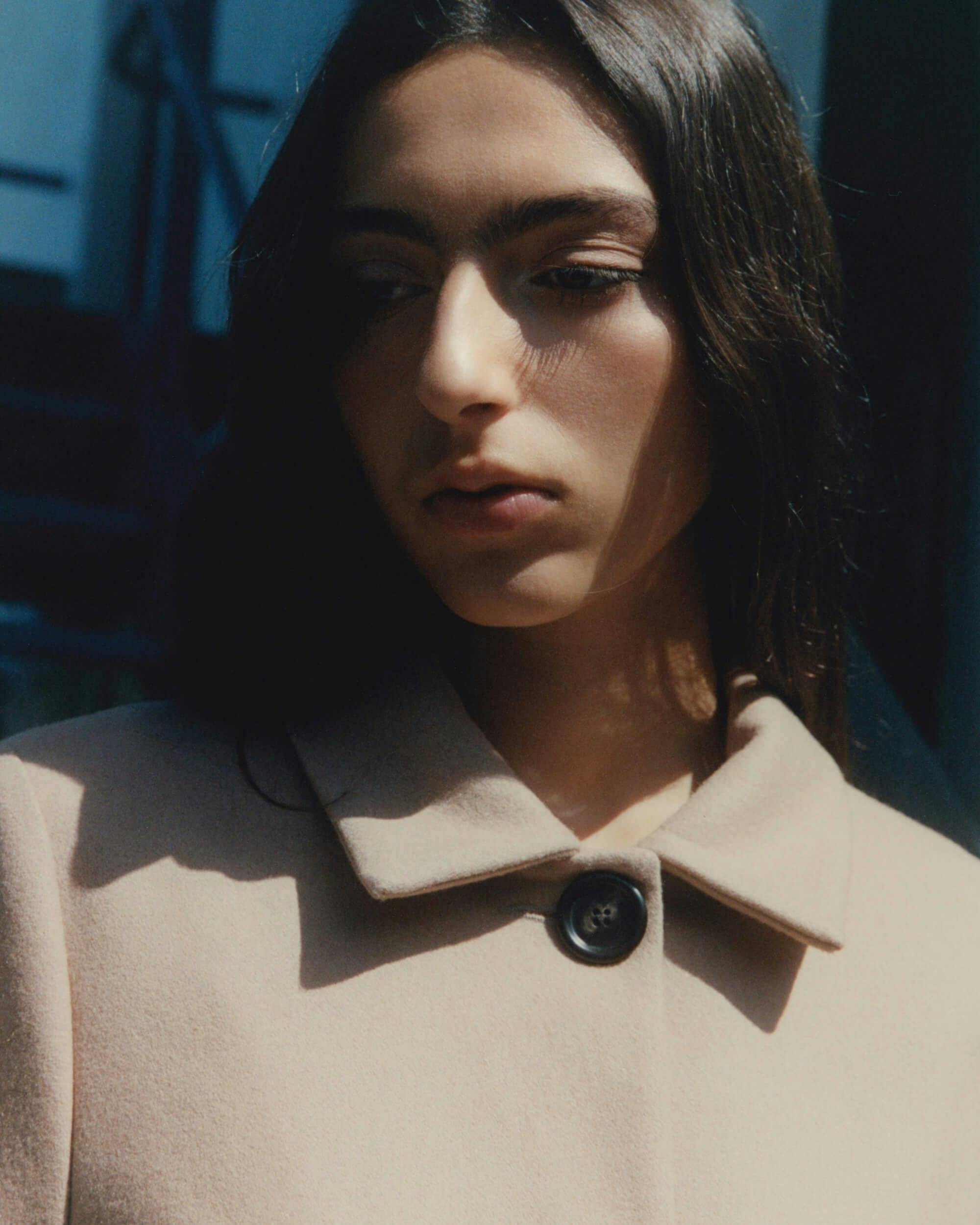 A woman with long dark hair wearing a beige coat with a large button stands outdoors in soft sunlight, looking down and slightly away from the camera. The background is blurred.