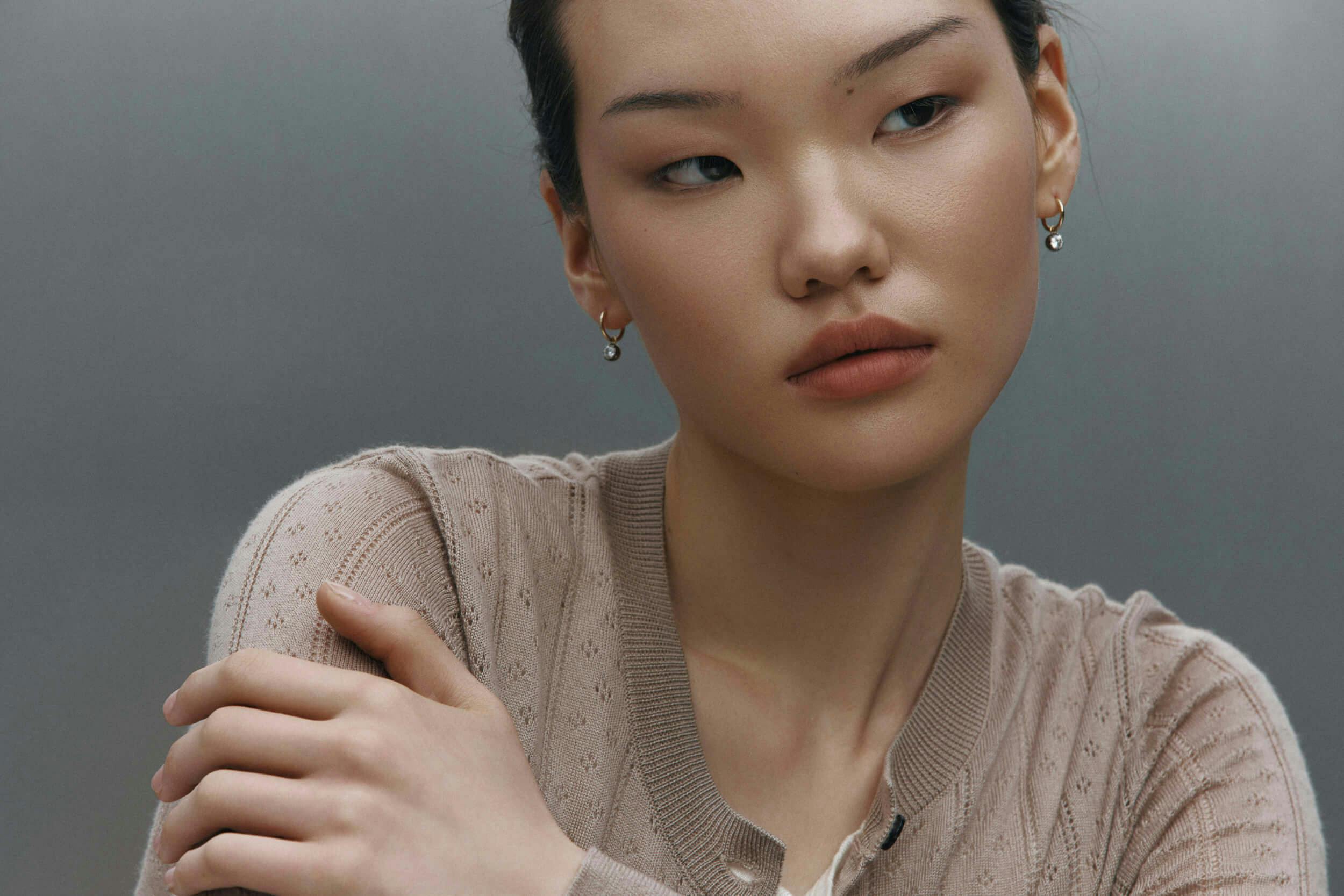 A young woman with fair skin and straight dark hair looks to the side with a neutral expression. She is wearing a light beige knit cardigan and small earrings, and her left arm crosses her body, touching her right shoulder.
