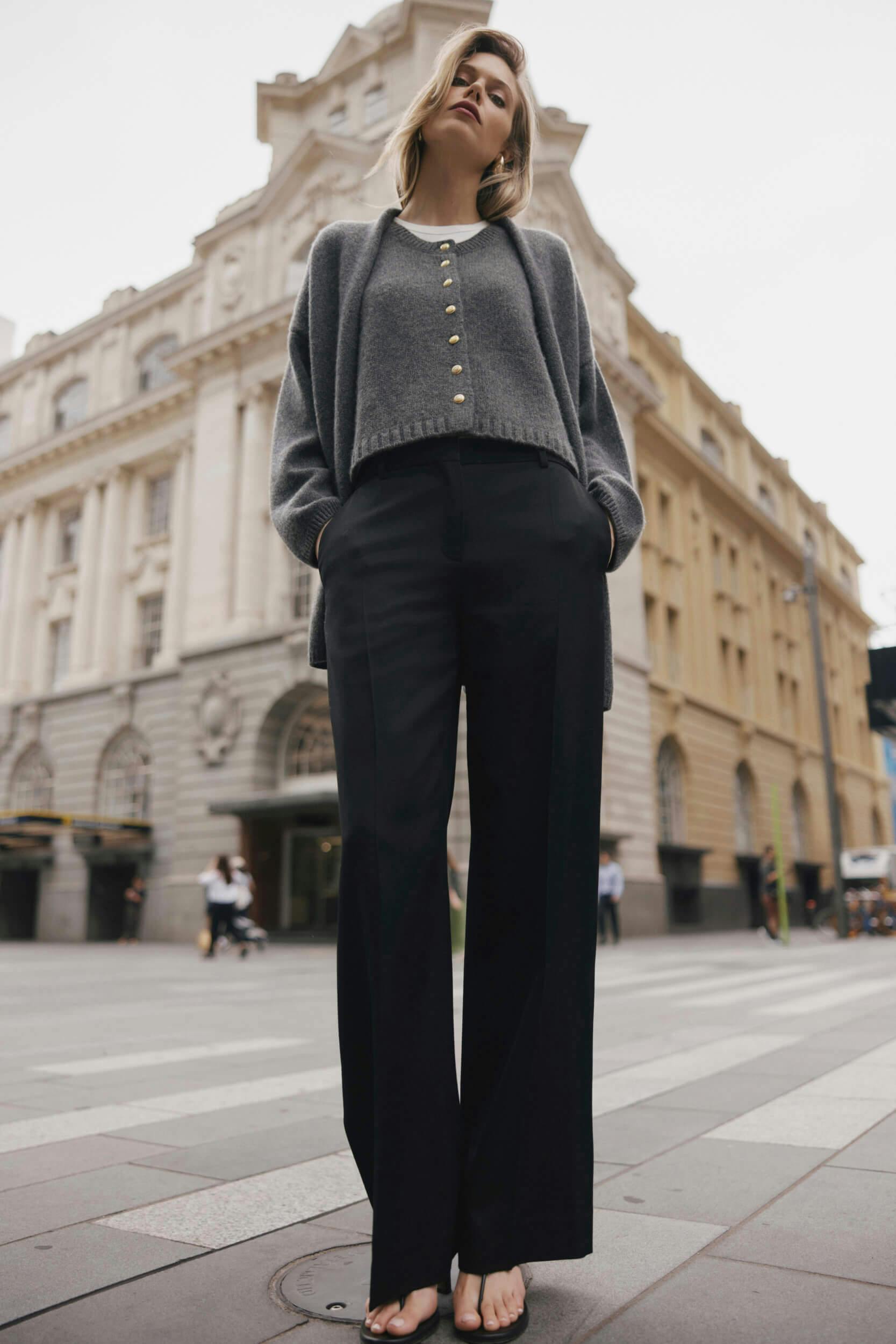 A person stands on a city street, wearing a grey cardigan with gold buttons, black wide-leg pants, and sandals. Their hands are in their pockets, and an ornate building is visible in the background.