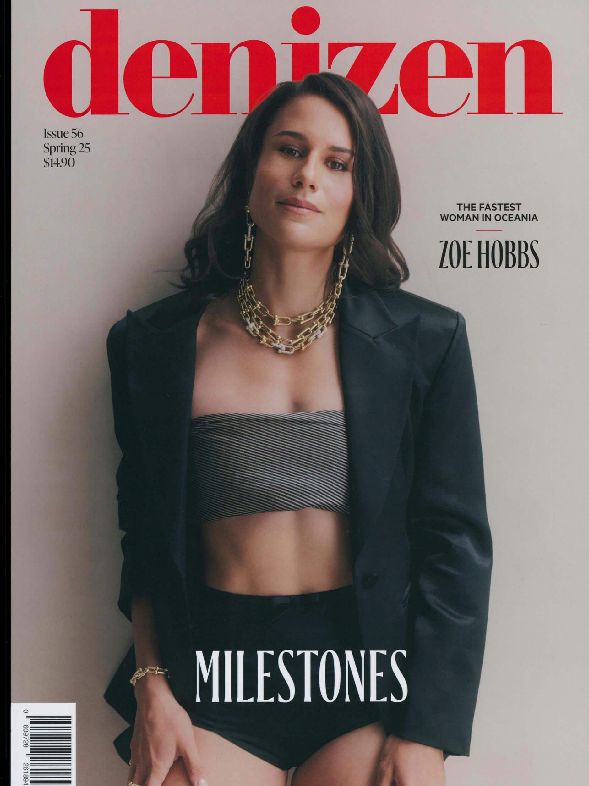 A woman stands against a plain background wearing a black blazer, a strapless striped crop top, and black shorts. The magazine cover headline reads 