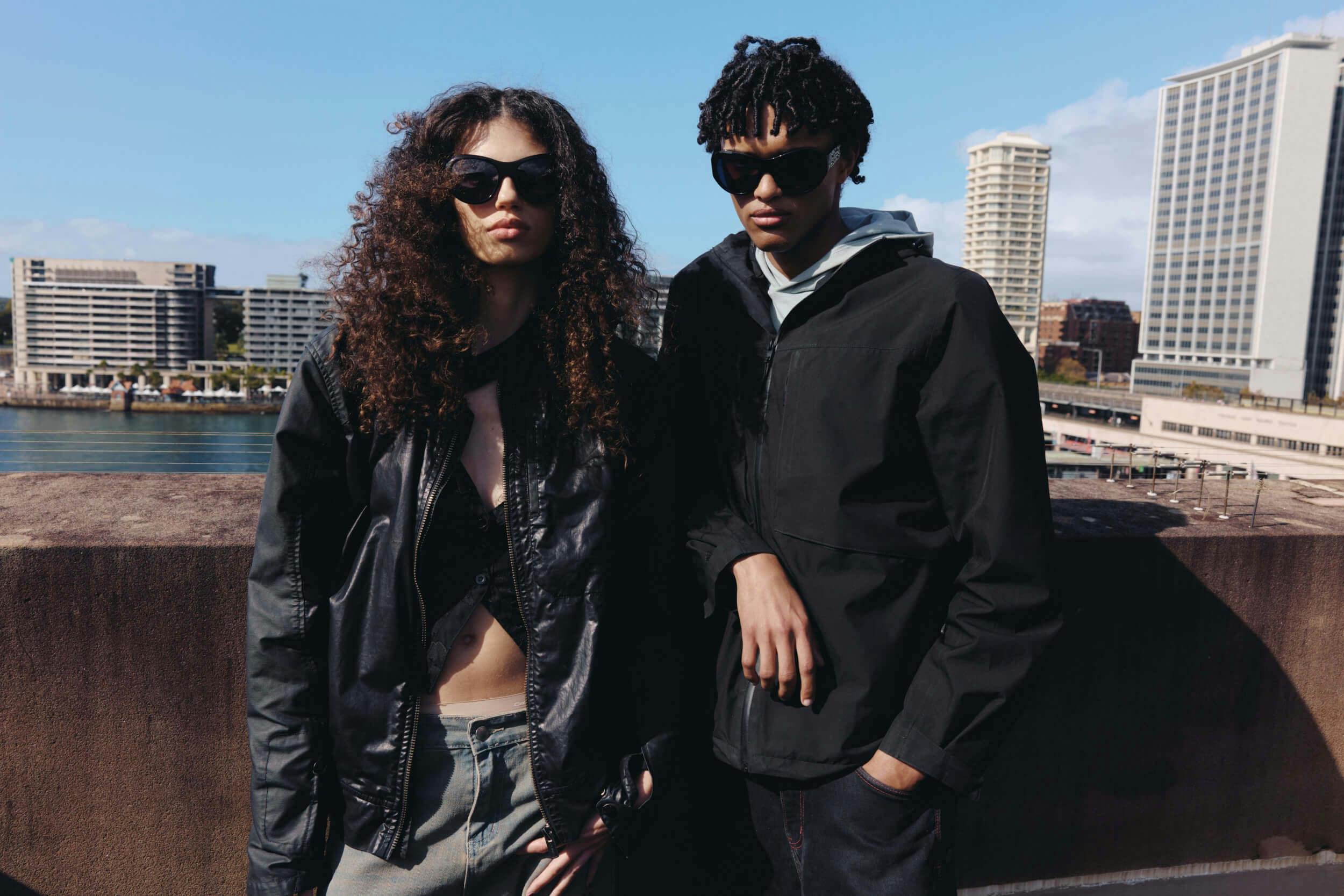 Two people wearing black jackets and sunglasses stand on a rooftop with a cityscape of tall buildings and blue sky in the background.