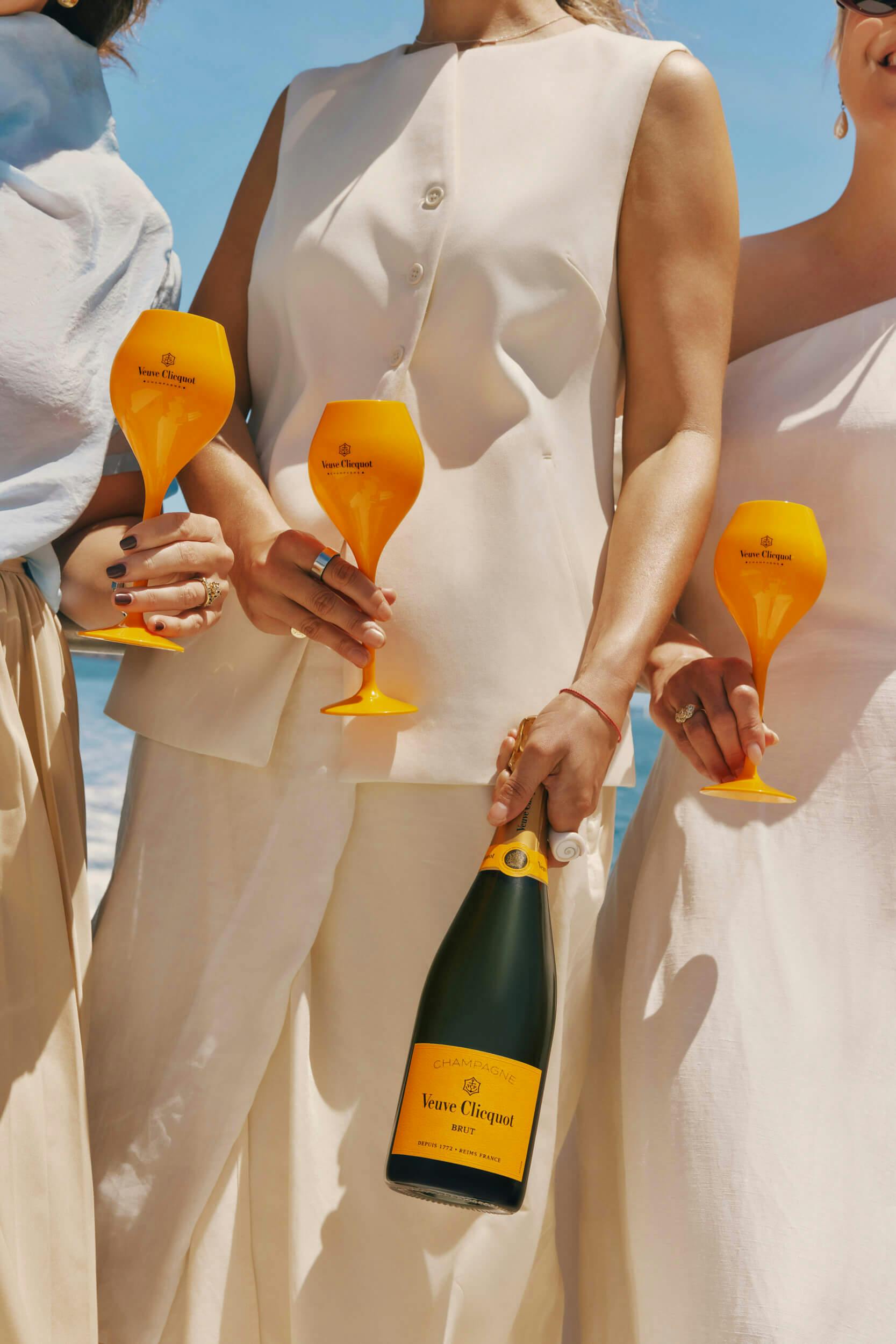 Three people in cream-colored outfits stand side by side, each holding a yellow Veuve Clicquot wine glass. One person holds a matching bottle of Veuve Clicquot champagne. The background shows a blue sky.
