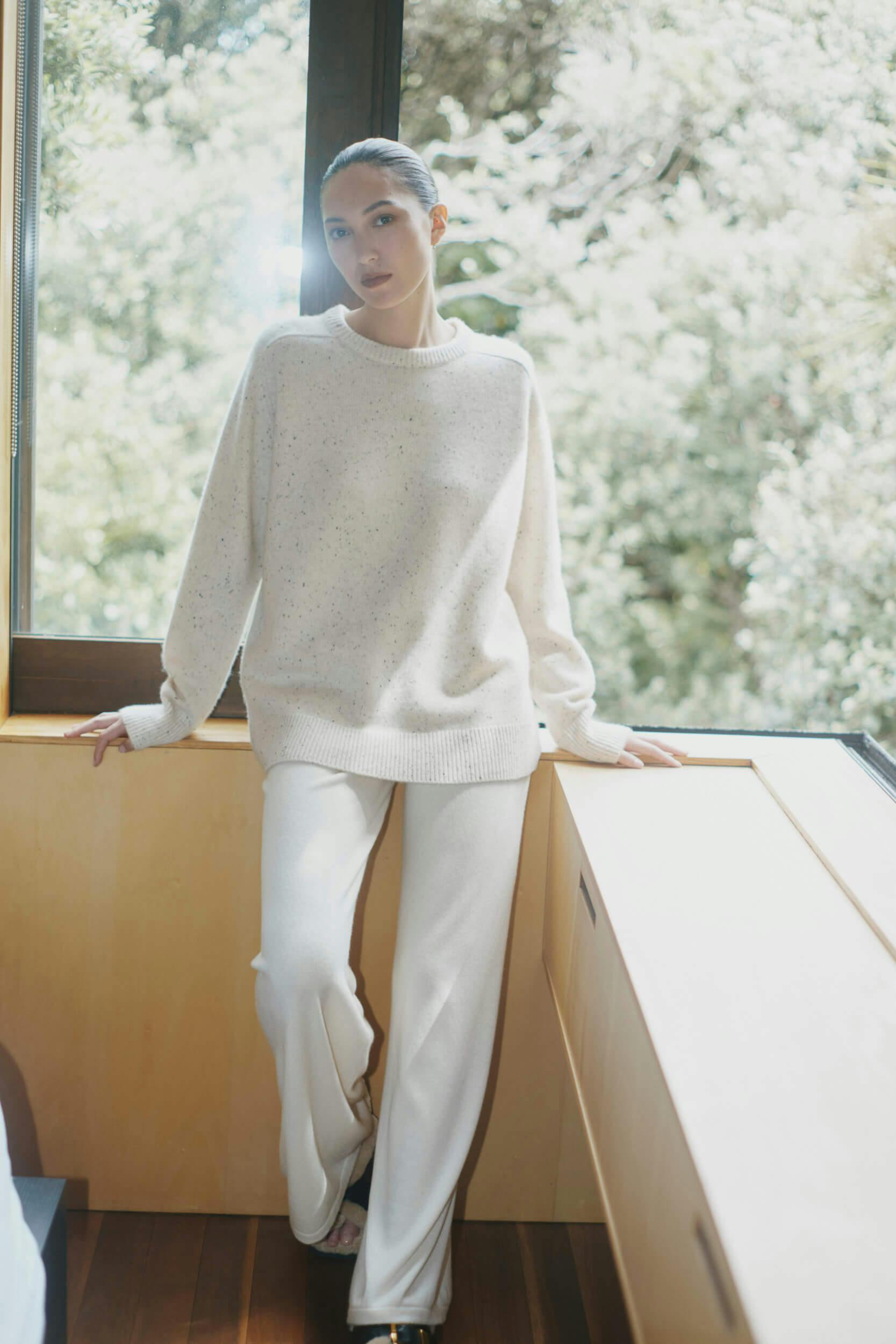 A person wearing a loose, cream-colored sweater and matching wide-legged pants is standing indoors by a window with trees visible outside. They are looking at the camera with one hand resting on a ledge.