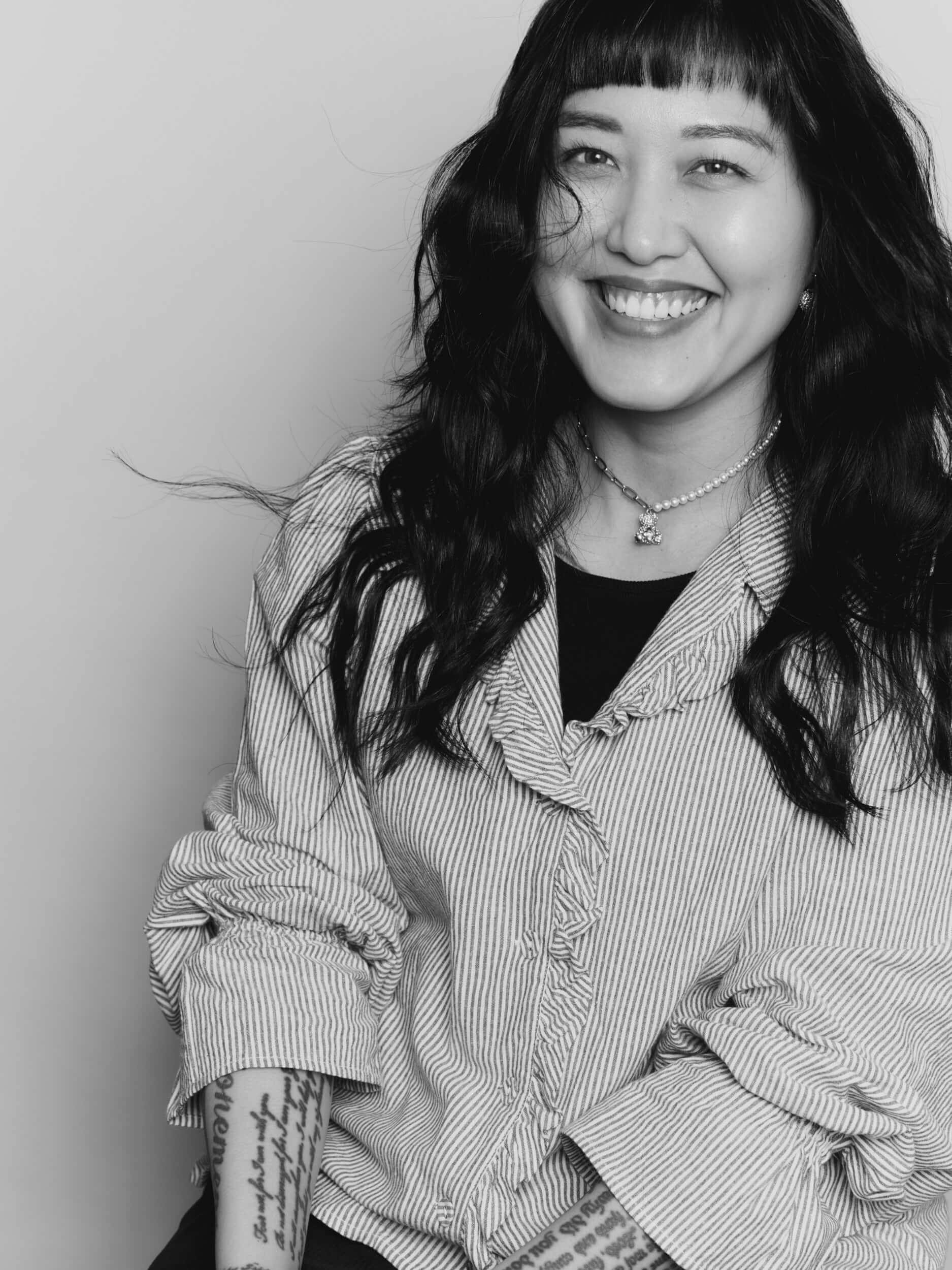 Black and white photo of a woman with long dark hair, wearing a striped shirt and a necklace, smiling at the camera. She has visible tattoos on her forearms and her hair is slightly tousled.