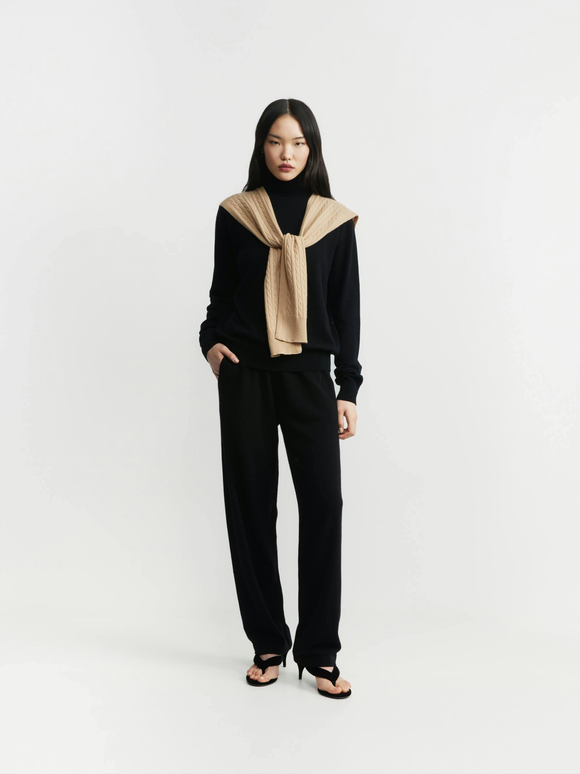 A woman stands against a plain white background, wearing a black turtleneck sweater, black trousers, black heeled sandals, and a tan sweater draped over her shoulders and tied in front. She has straight, long hair.