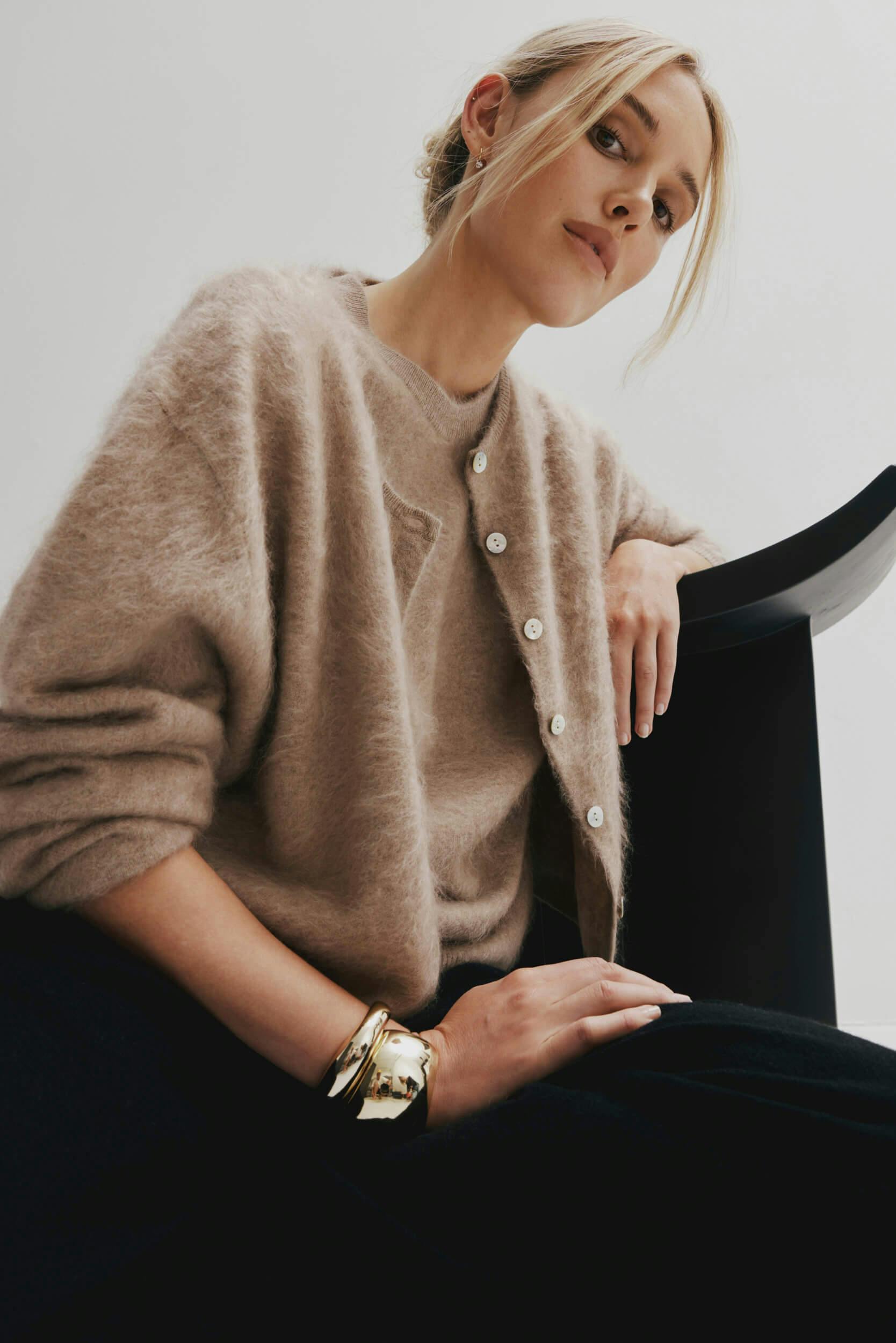 A person with blonde hair tied back is seated, wearing a beige fuzzy cardigan, a neutral top, black pants, and gold bangles, with one arm resting on a black curved chair.