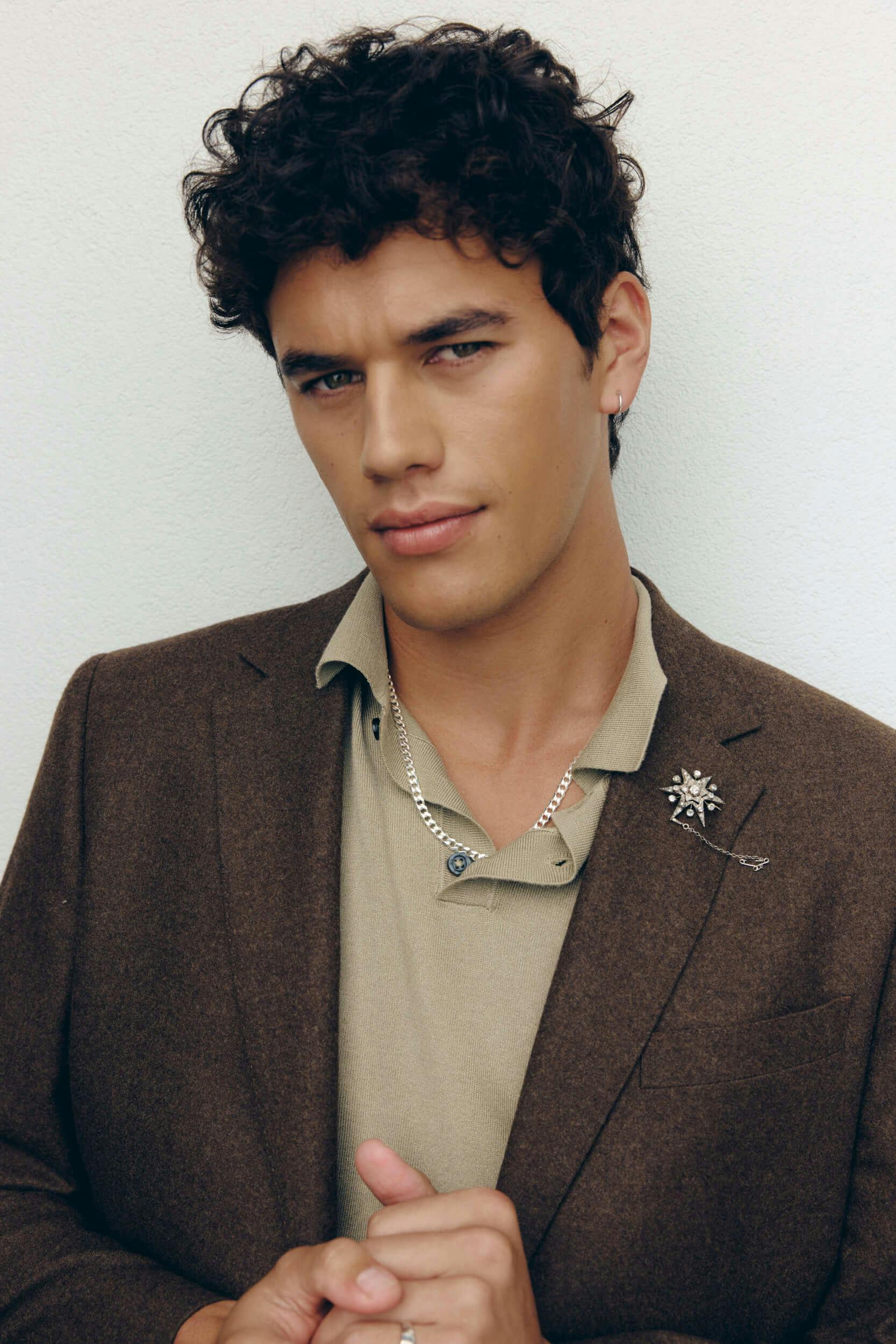 A person with short curly hair wearing a brown blazer over a beige shirt, accessorized with a chain necklace and a brooch, stands against a plain light-colored background.