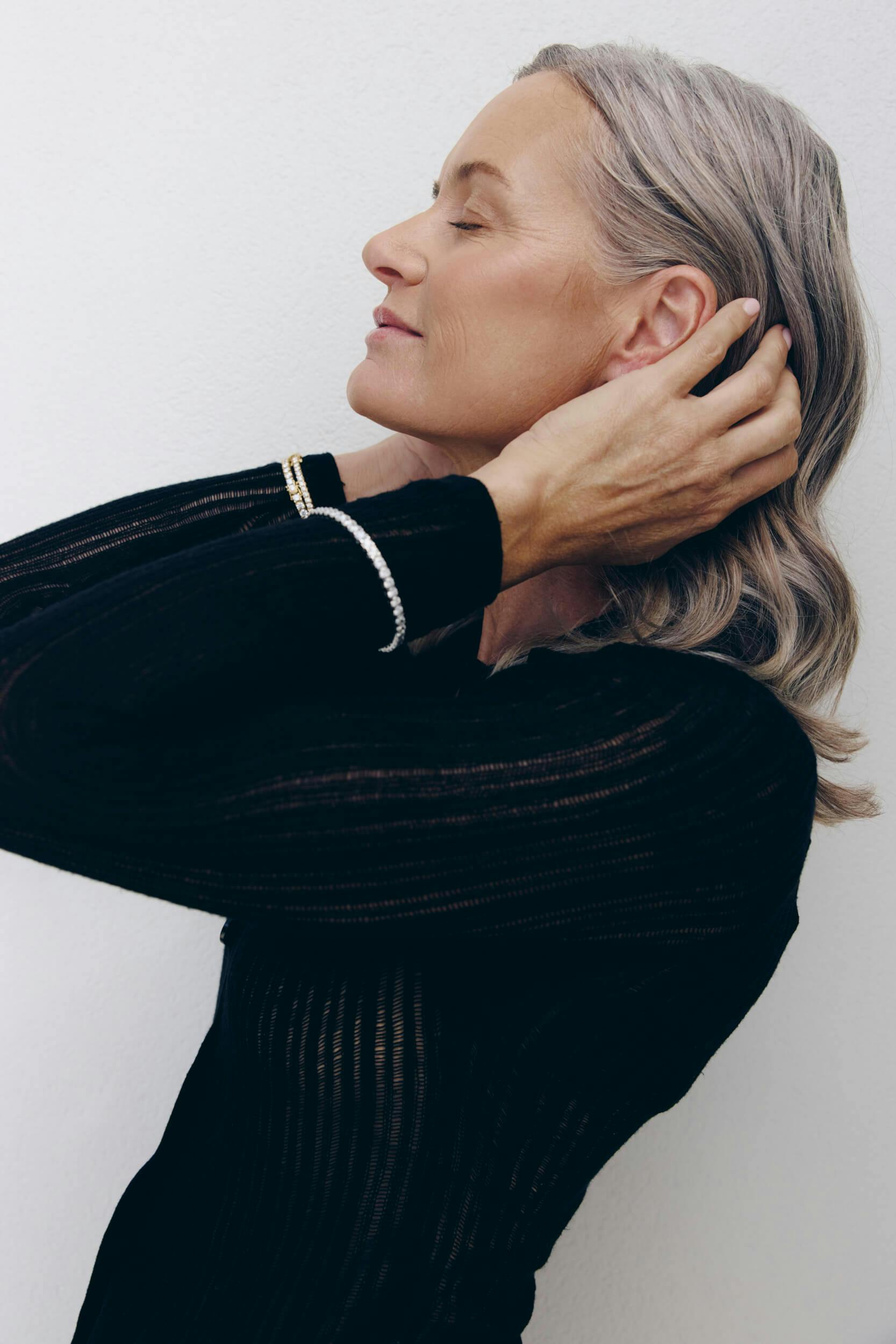 A woman with gray hair, wearing a black long-sleeve top and a bracelet, stands in profile against a white background with her eyes closed and one hand touching her hair.