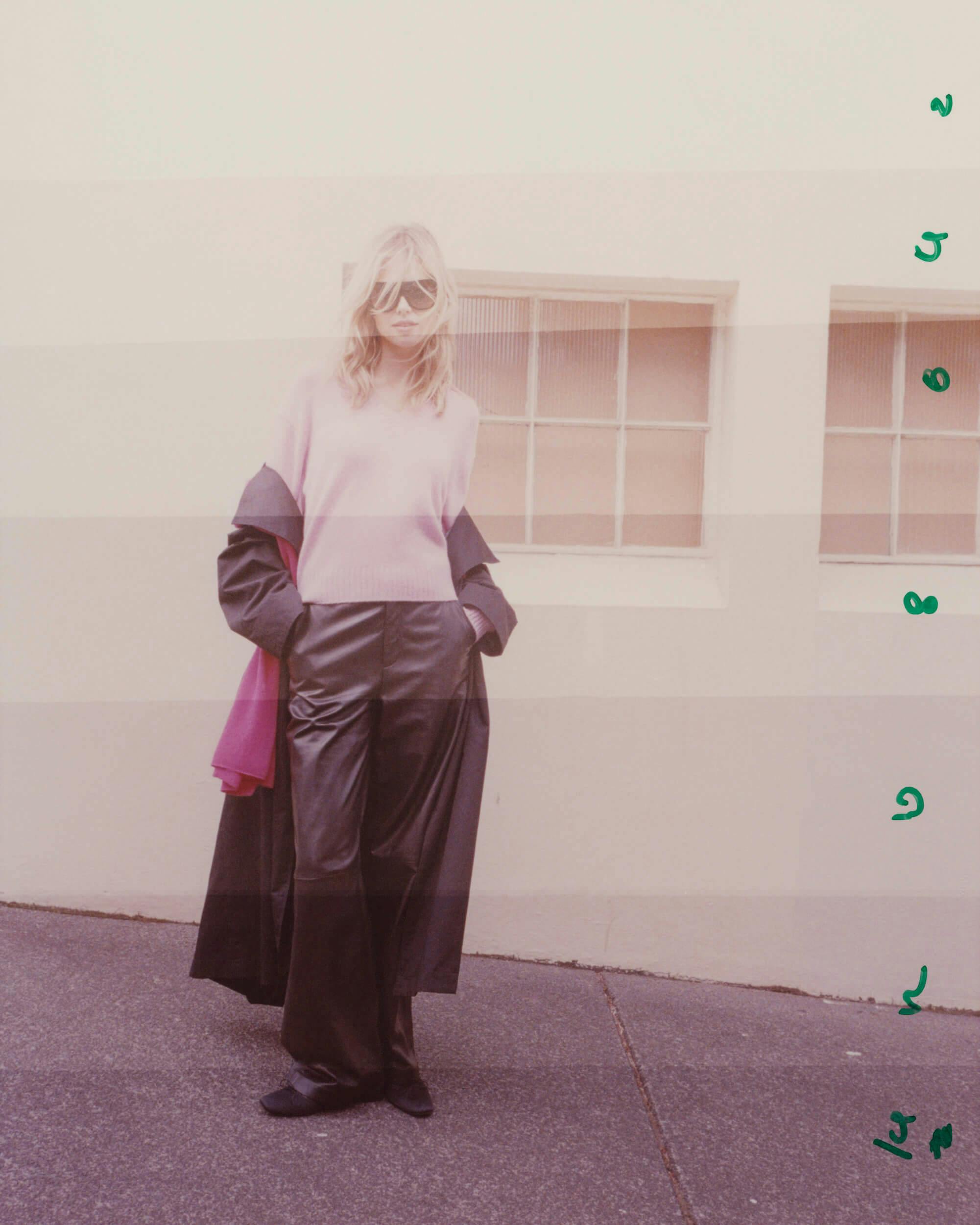 A person wearing sunglasses, a pink sweater, black pants, and a long black coat stands on a paved sidewalk in front of a cream-colored wall with two windows. Green numbers or letters appear vertically on the right side.