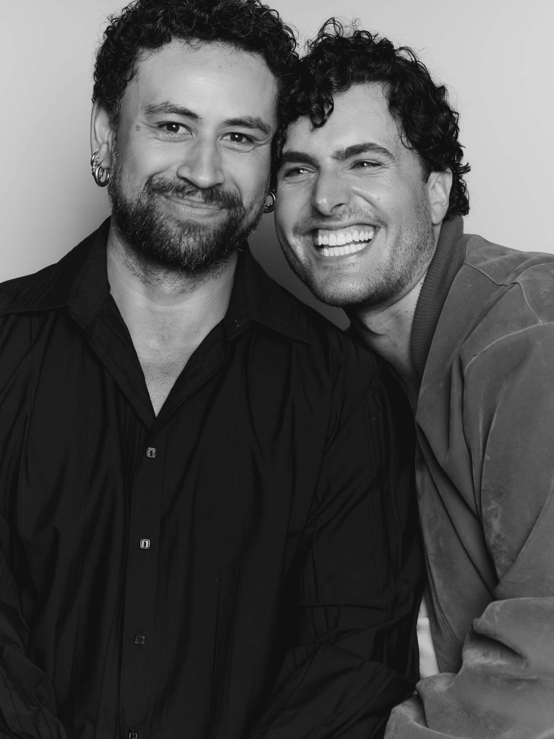 Two people pose closely together, smiling. The person on the left has short curly hair, a beard, and is wearing a dark shirt. The person on the right has curly hair, is clean-shaven, and is wearing a light-colored jacket. The image is in black and white.