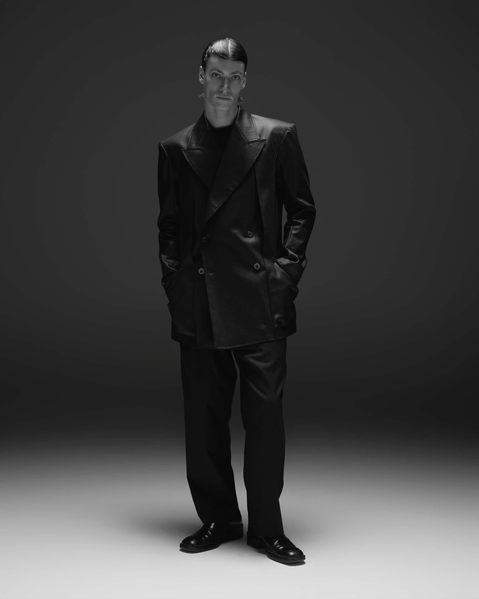 A person stands with hands in pockets, wearing a double-breasted dark blazer, matching trousers, and dress shoes. The background is plain and dark, and the lighting is focused on the subject, creating a dramatic effect.