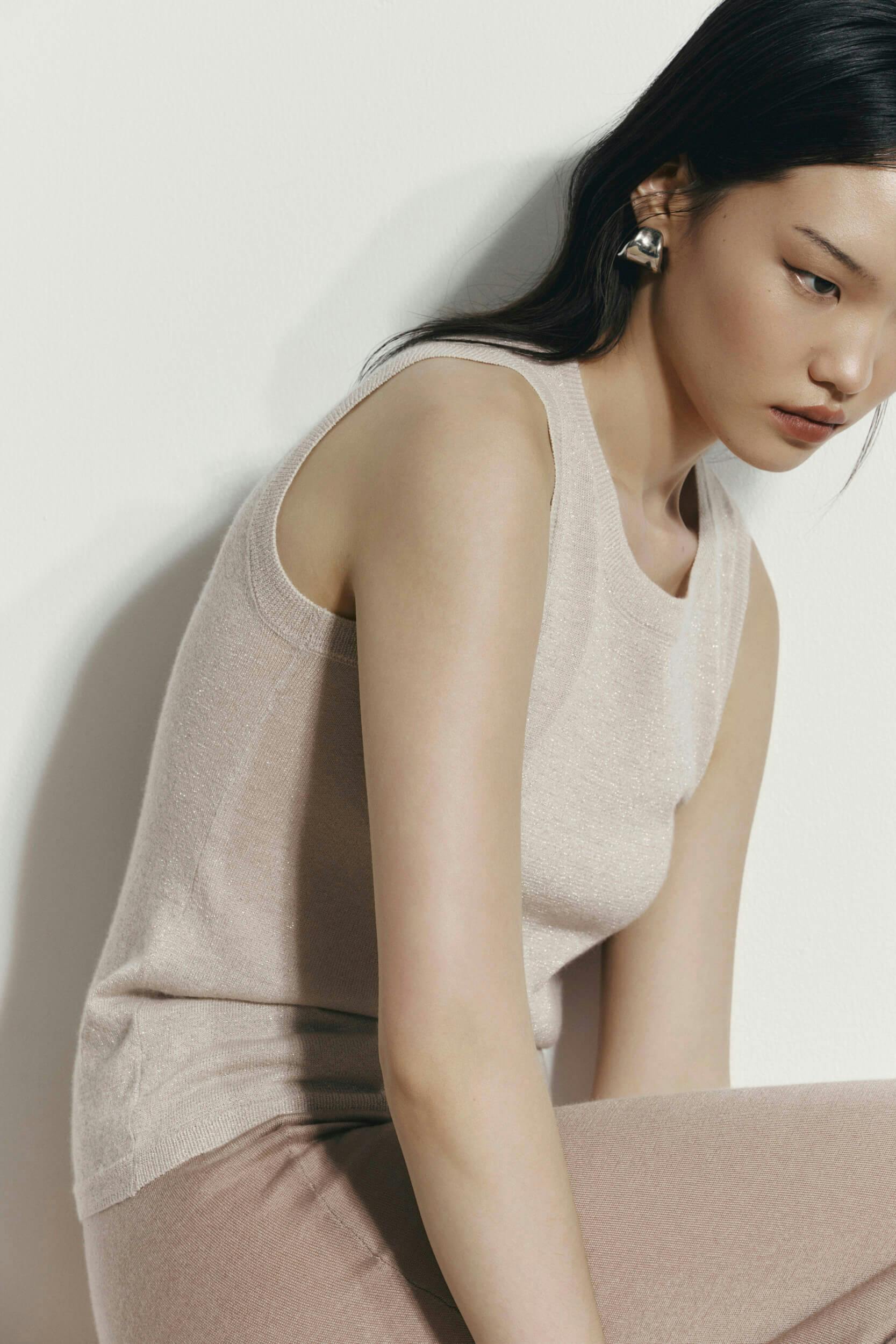 A person with long dark hair wears a sleeveless beige top and beige pants, sitting against a white background and looking down with a neutral expression.