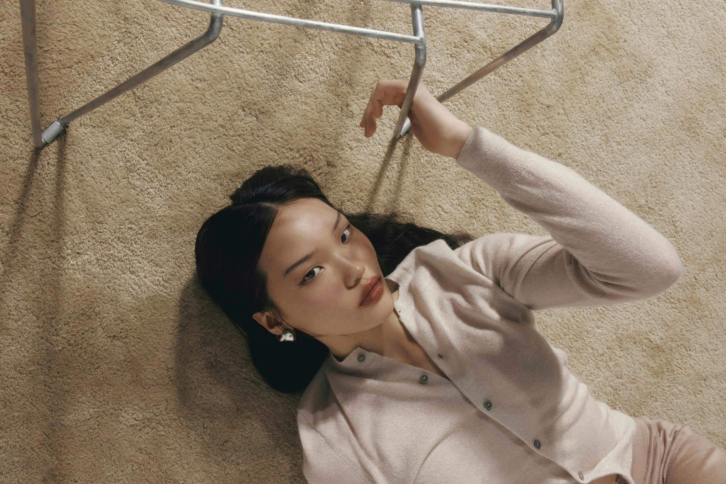 A woman with long dark hair lies on a beige carpet, wearing a light pink buttoned cardigan. She looks at the camera and raises one arm to hold a metal clothes rack above her.