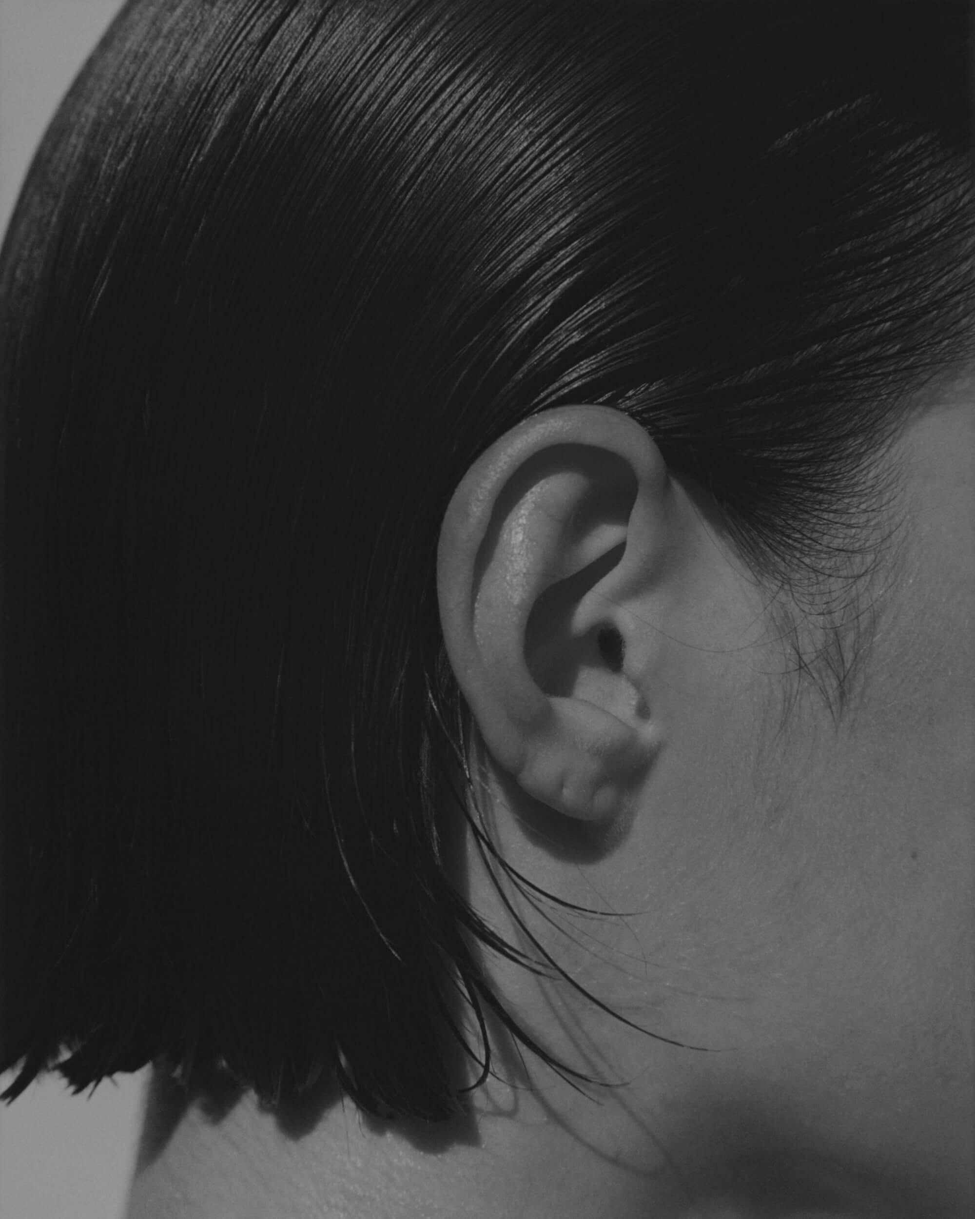 Close-up black and white photo of the side of a person's head, focusing on the ear and short, straight dark hair that is tucked behind the ear. The skin and hair texture are clearly visible.