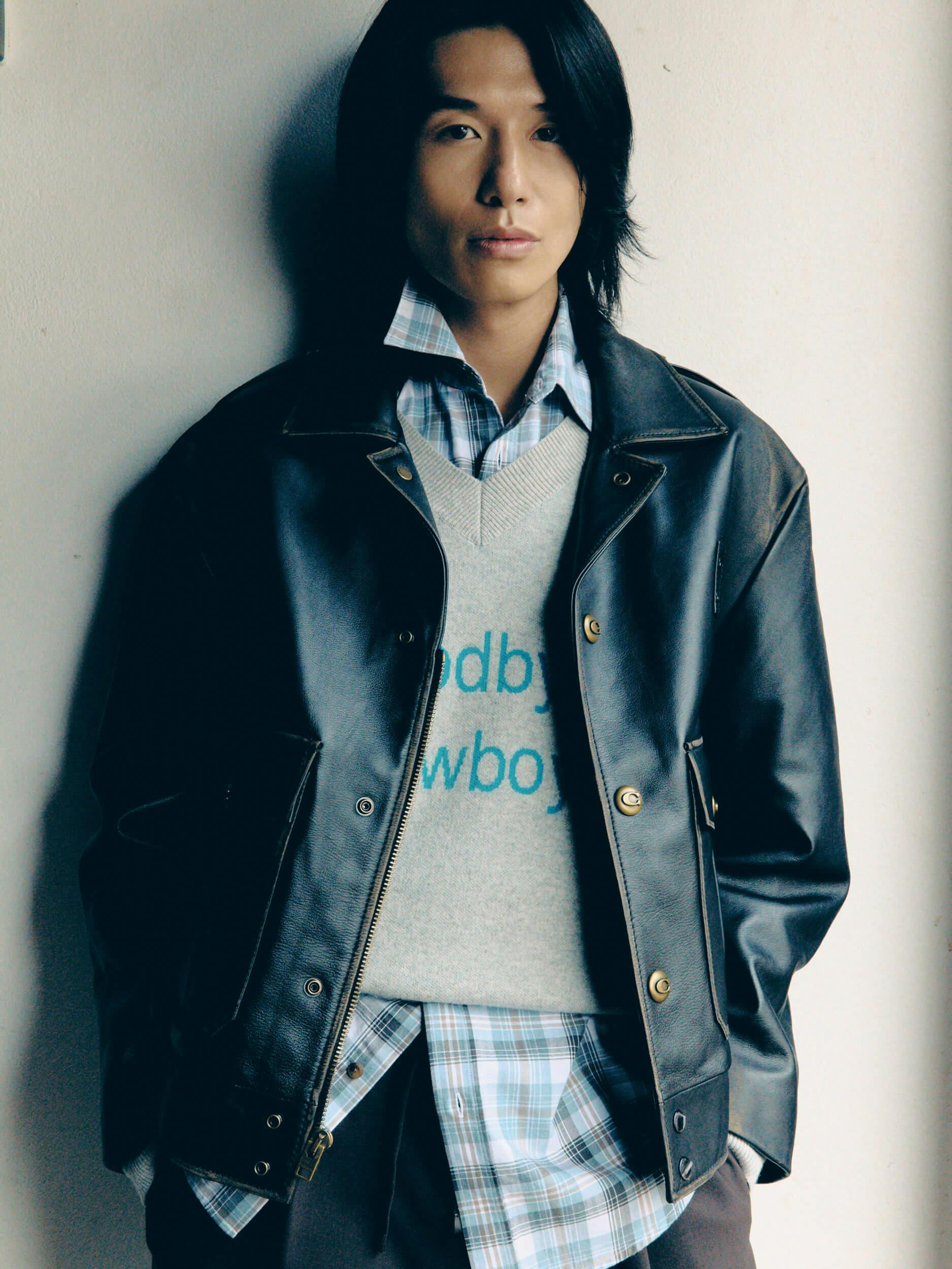 A person with long dark hair leans against a white wall, wearing a black leather jacket over a plaid shirt and a light gray sweater with blue text partially reading 