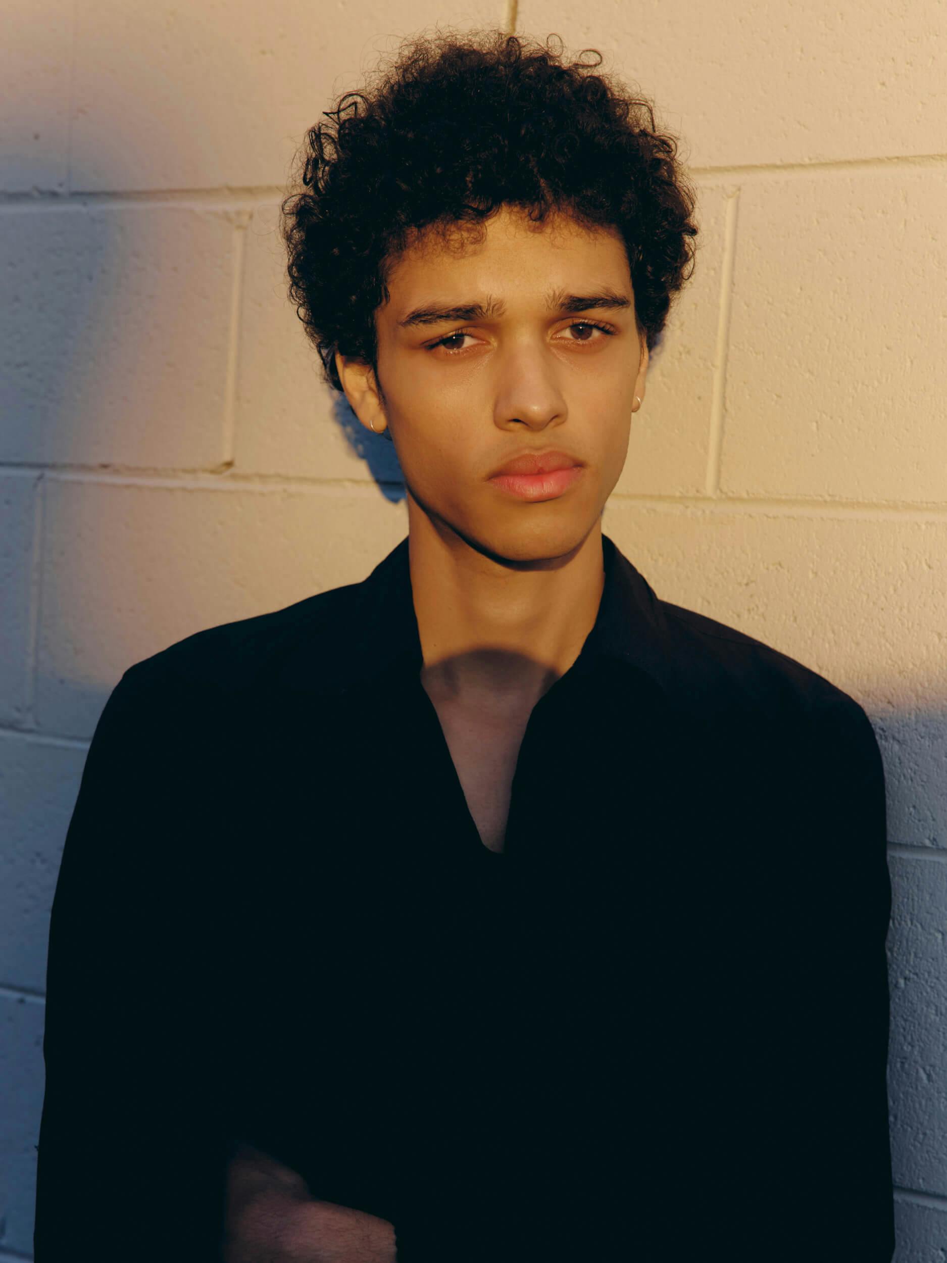 A person with short curly hair and a serious expression stands in front of a light-colored brick wall, wearing a black shirt. The lighting creates shadows on the wall and across the person’s face.