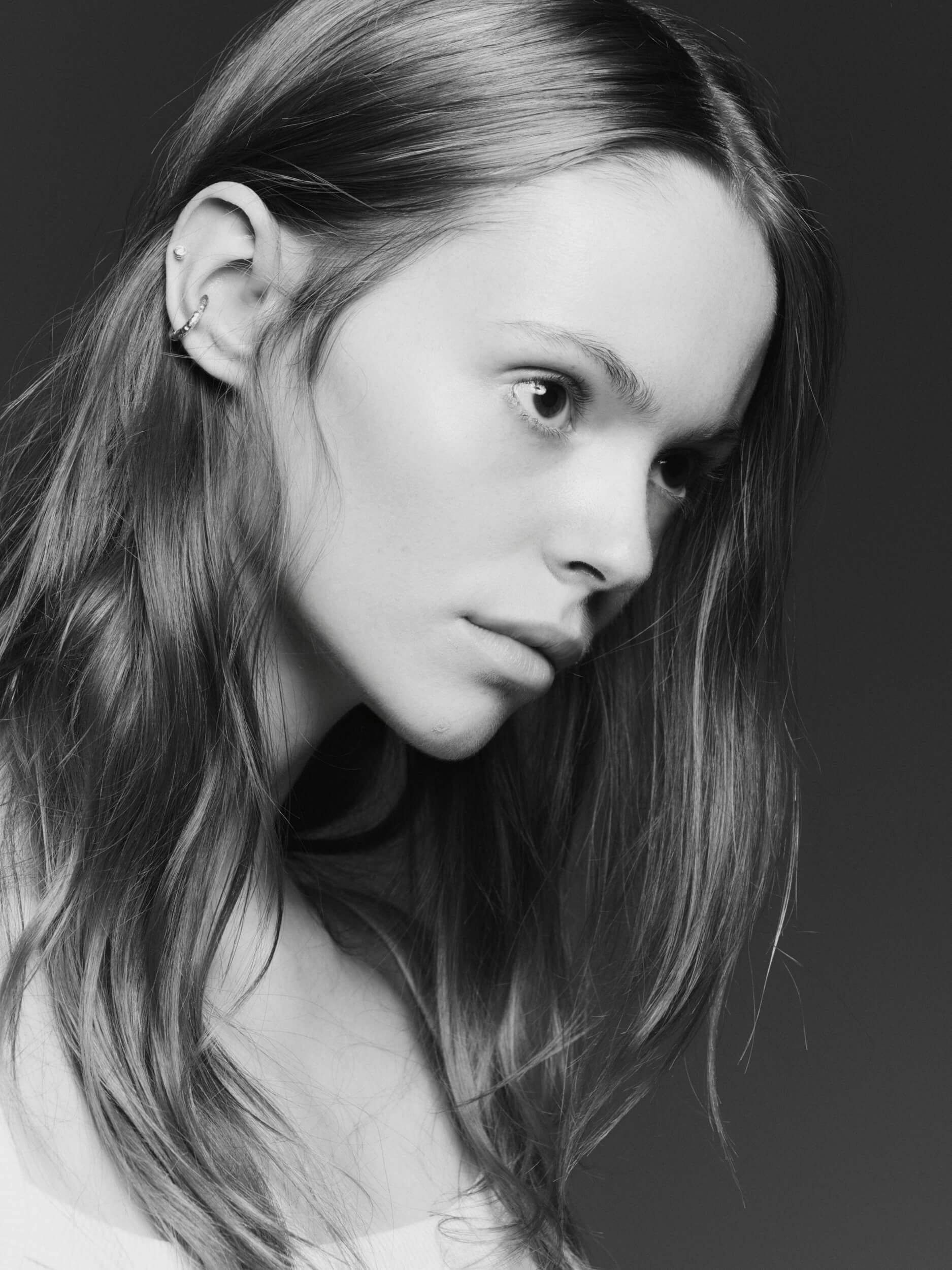 A young person with long, straight hair and multiple earrings in their left ear looks slightly downward against a dark background. The image is in black and white.