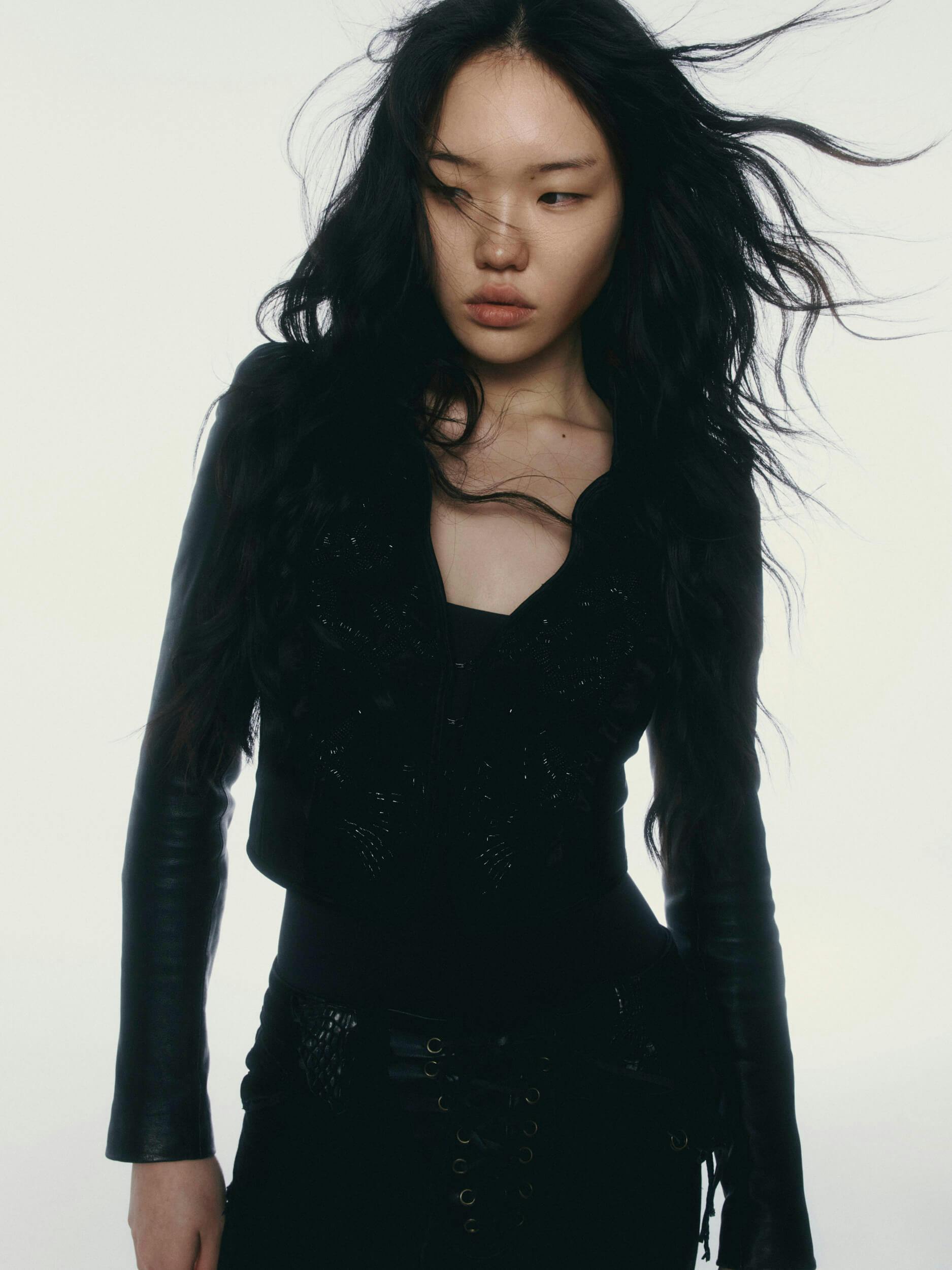A woman with long, wavy black hair wearing a fitted black jacket and black pants stands against a plain, light background. Her hair is blown by the wind, and she looks slightly downward.