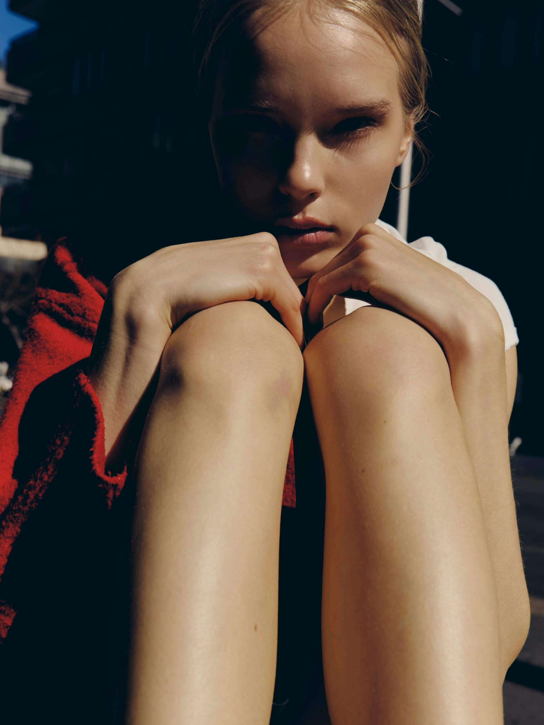 A person sits outdoors in bright sunlight with knees pulled to their chest, hands clasped over their knees, and a serious facial expression. Shadows partially cover their face and legs.