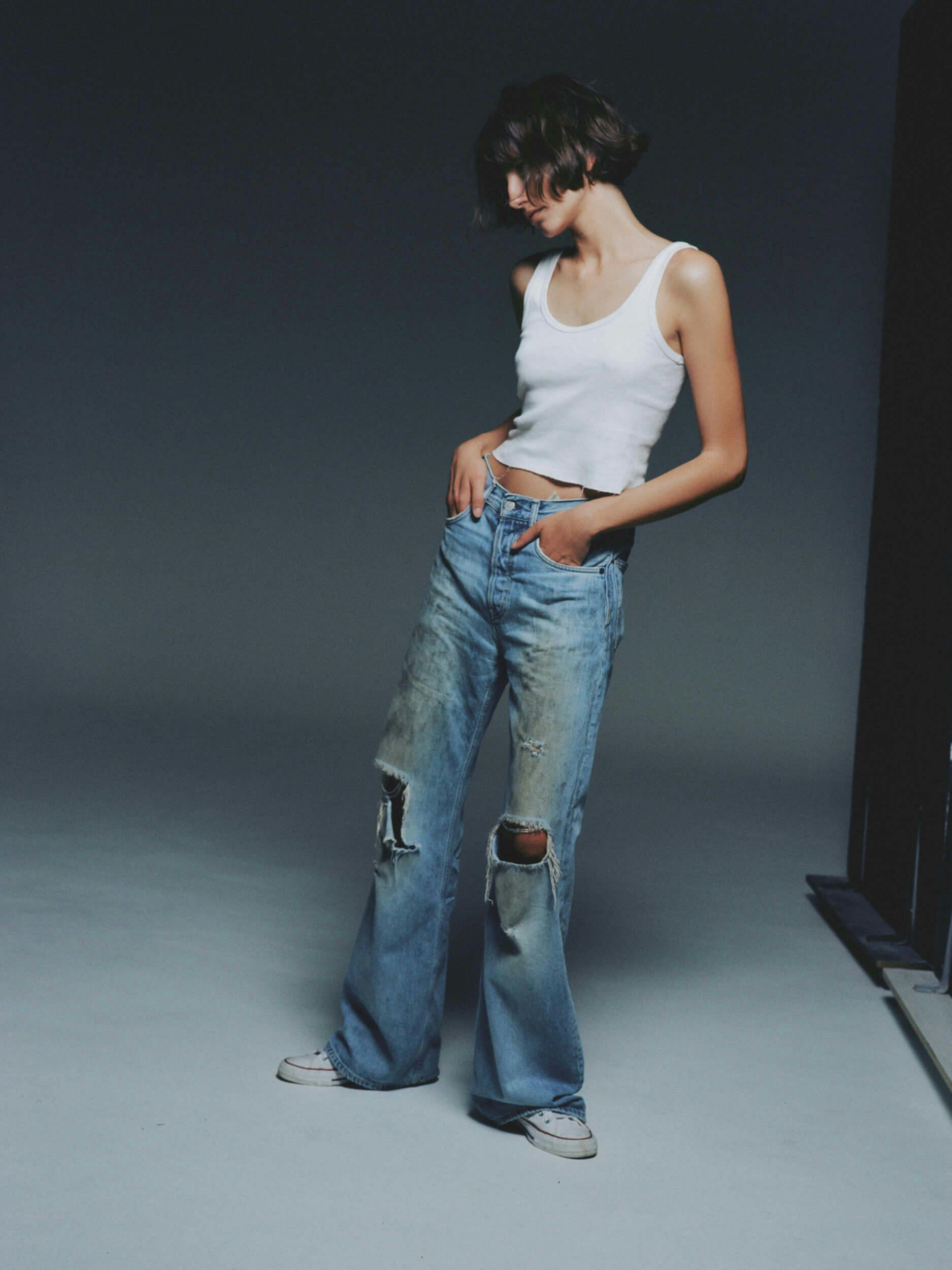 A person stands against a dark background wearing a white tank top, distressed blue jeans with knee holes, and white sneakers. They have short hair and are looking down with one hand in a pocket.