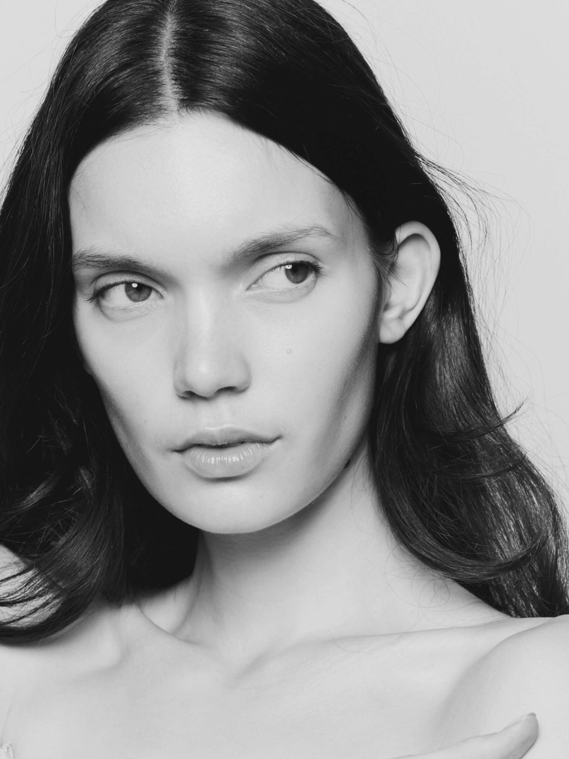 A black and white close-up photo of a woman with straight, dark hair, parted in the middle, looking slightly to the side with a neutral expression against a plain background.