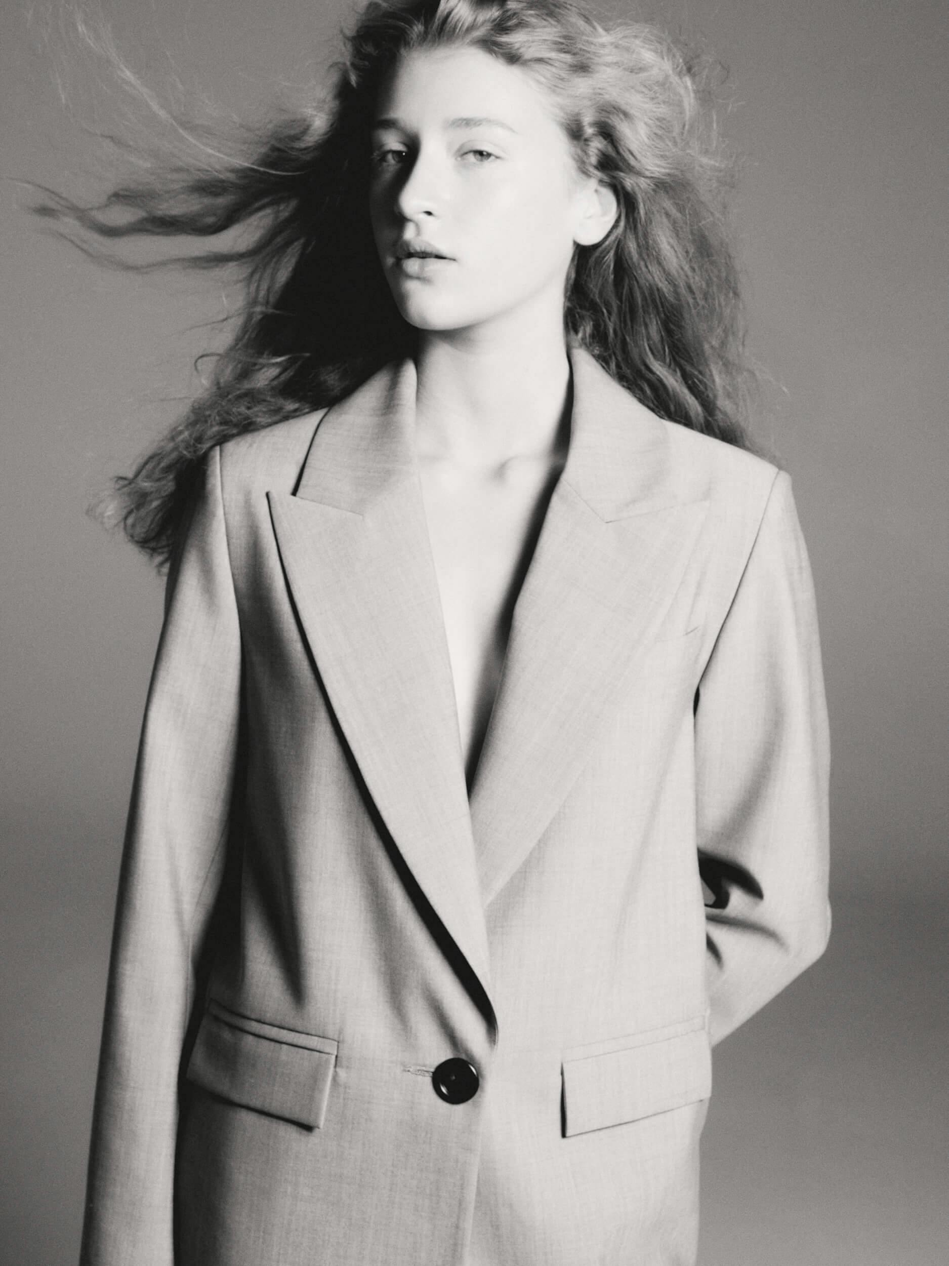 Black and white photo of a person with long, wavy hair wearing an oversized blazer. The person is looking slightly to the side with a neutral expression, and their hair appears to be blowing in the wind.