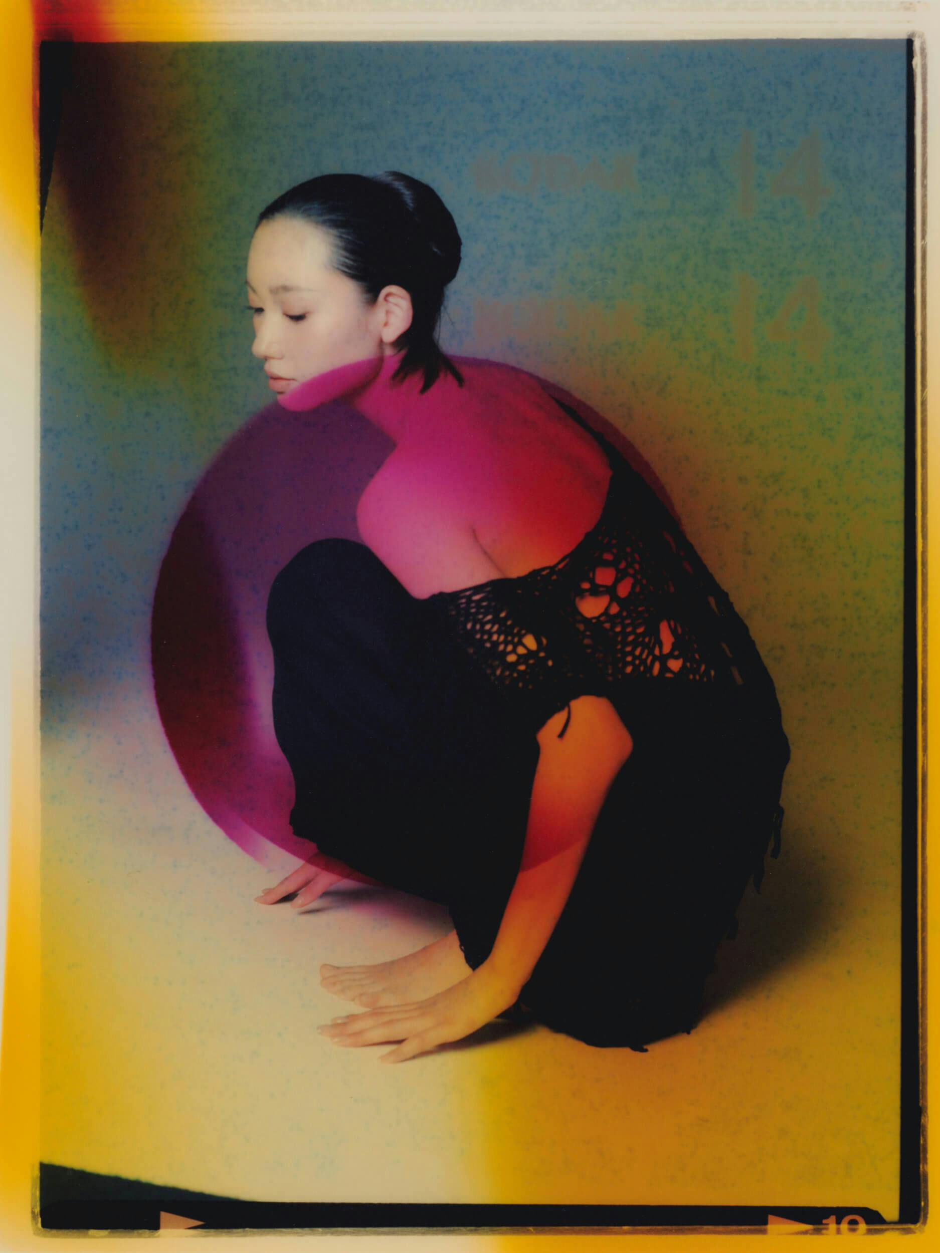 A person with dark hair slicked back crouches beside a large pink sphere, wearing loose black pants and a black crochet top, with dramatic colored lighting creating purple, yellow, and blue hues on the background and skin.