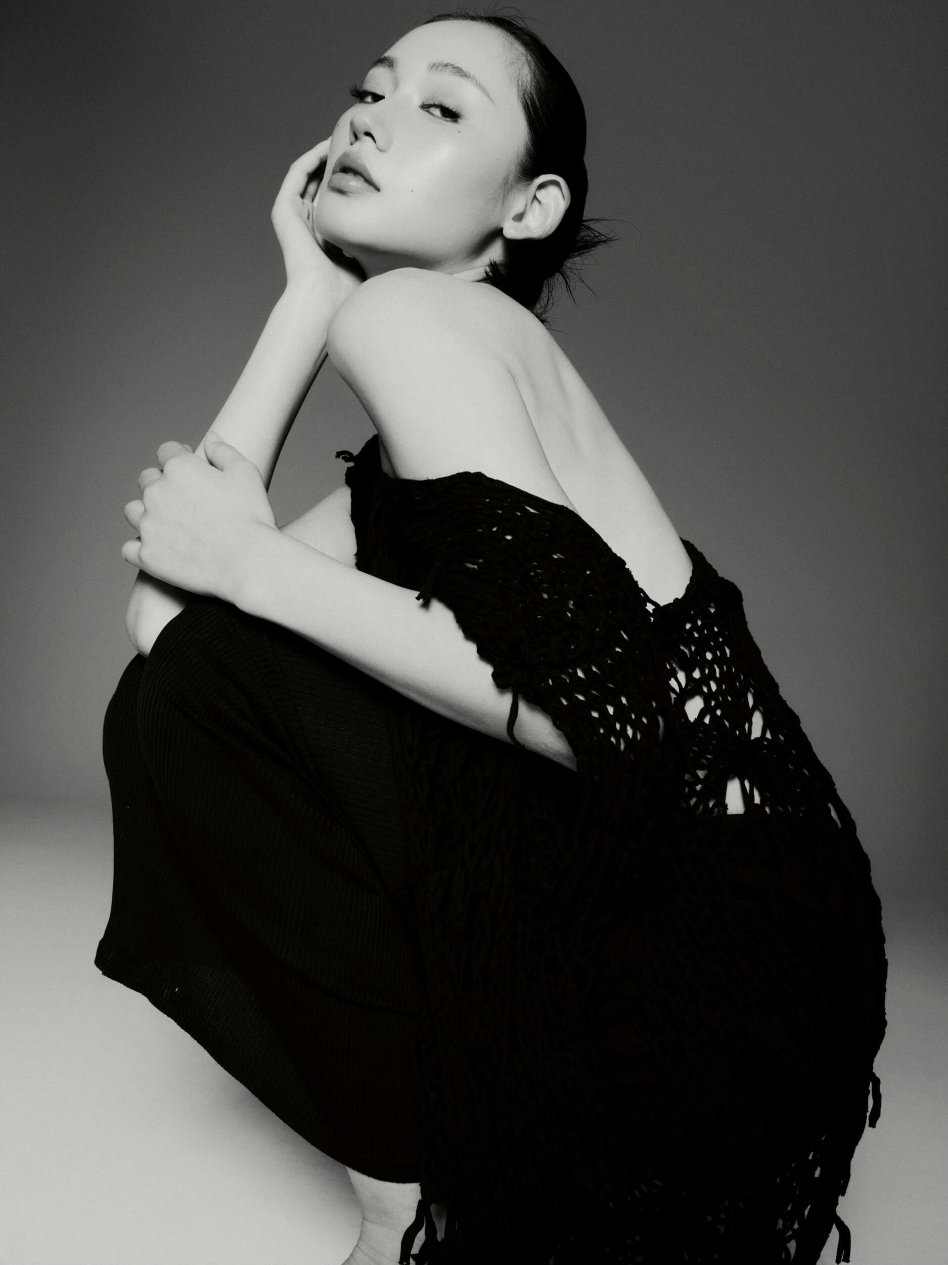 A person with dark hair pulled back is squatting and looking at the camera. They wear a loosely knit, off-the-shoulder black garment and dark pants in a studio with plain background. The image is black and white.