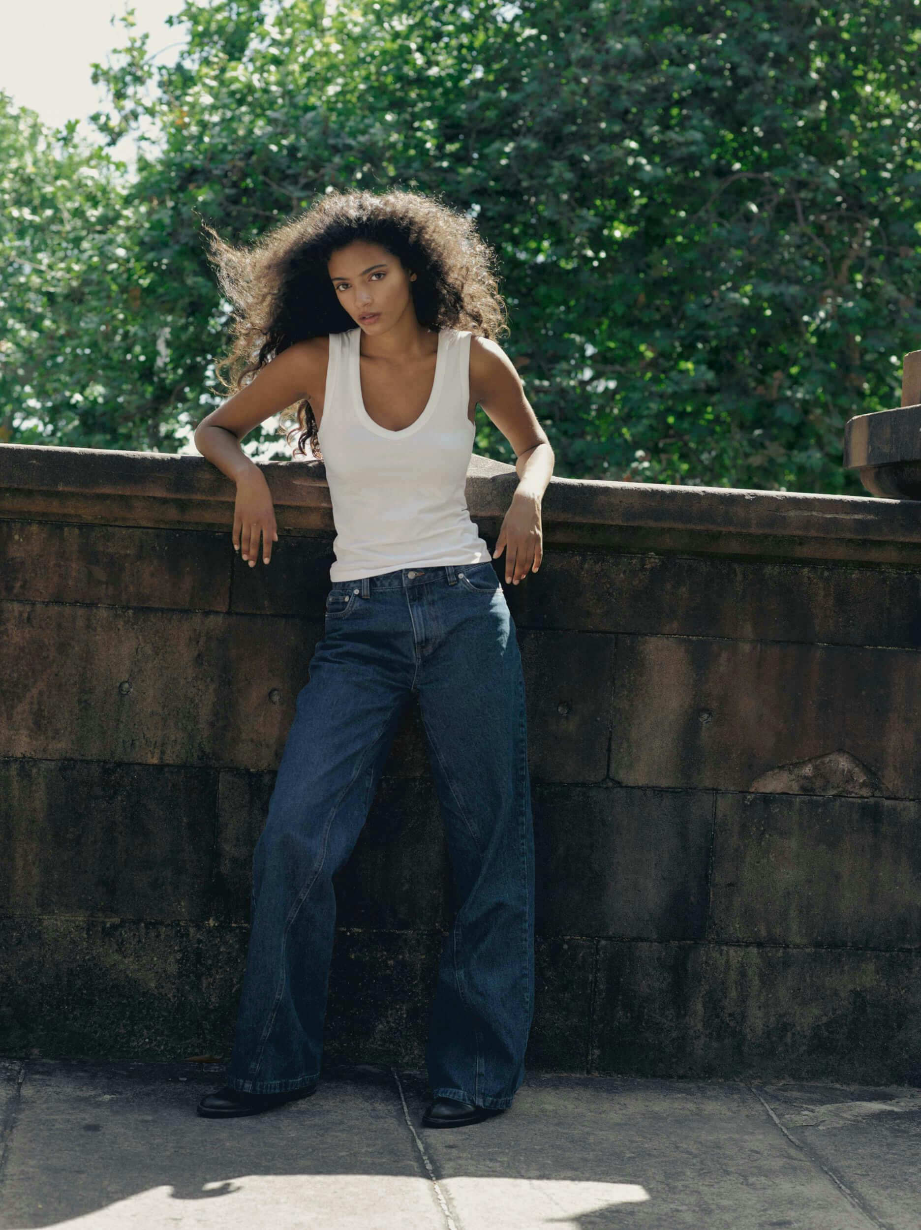 A person with long curly hair stands outdoors in front of a stone wall, wearing a white sleeveless top and loose-fitting blue jeans, with trees visible in the background.