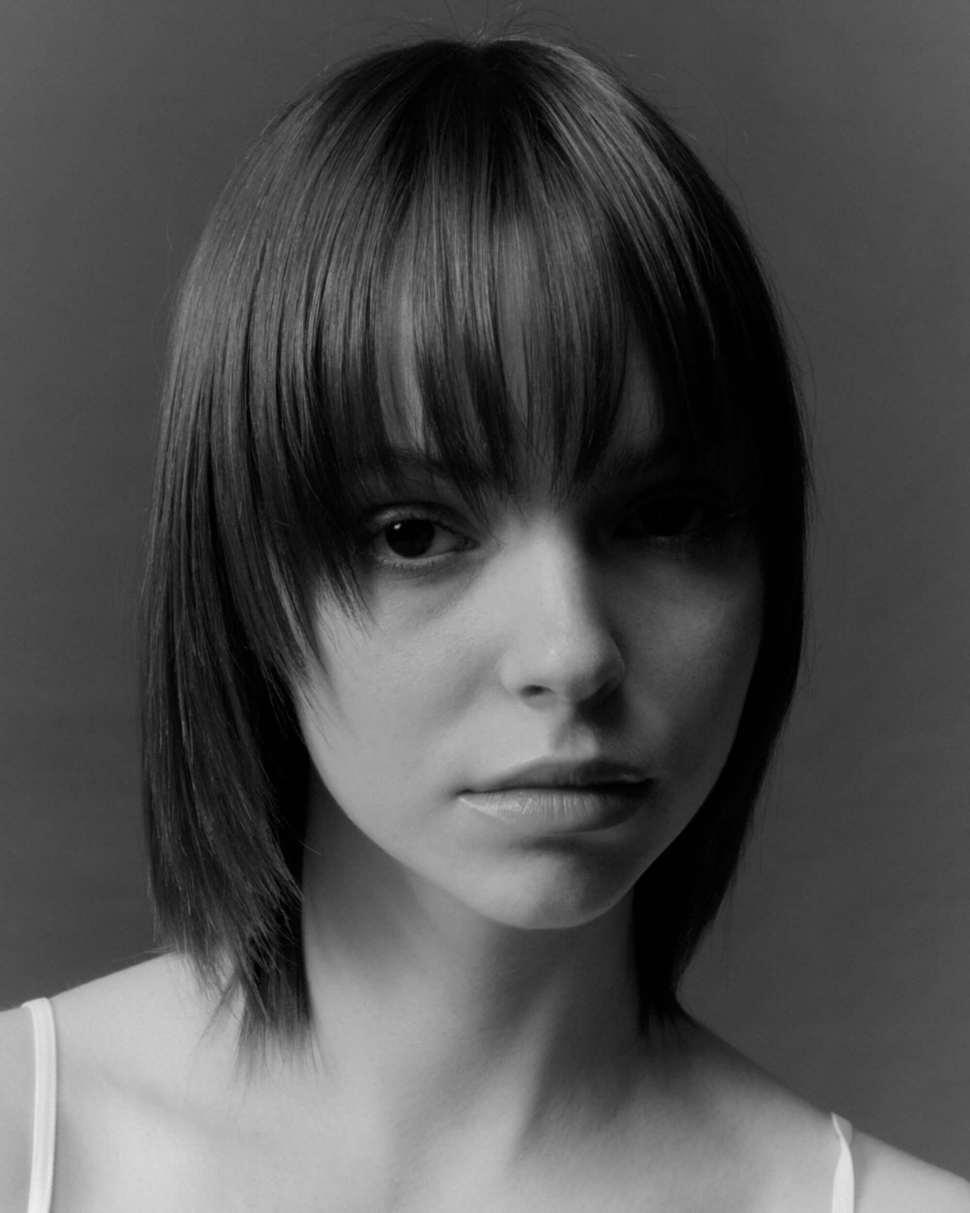A young person with straight, medium-length hair and bangs looks at the camera with a neutral expression against a plain background. The image is in black and white.