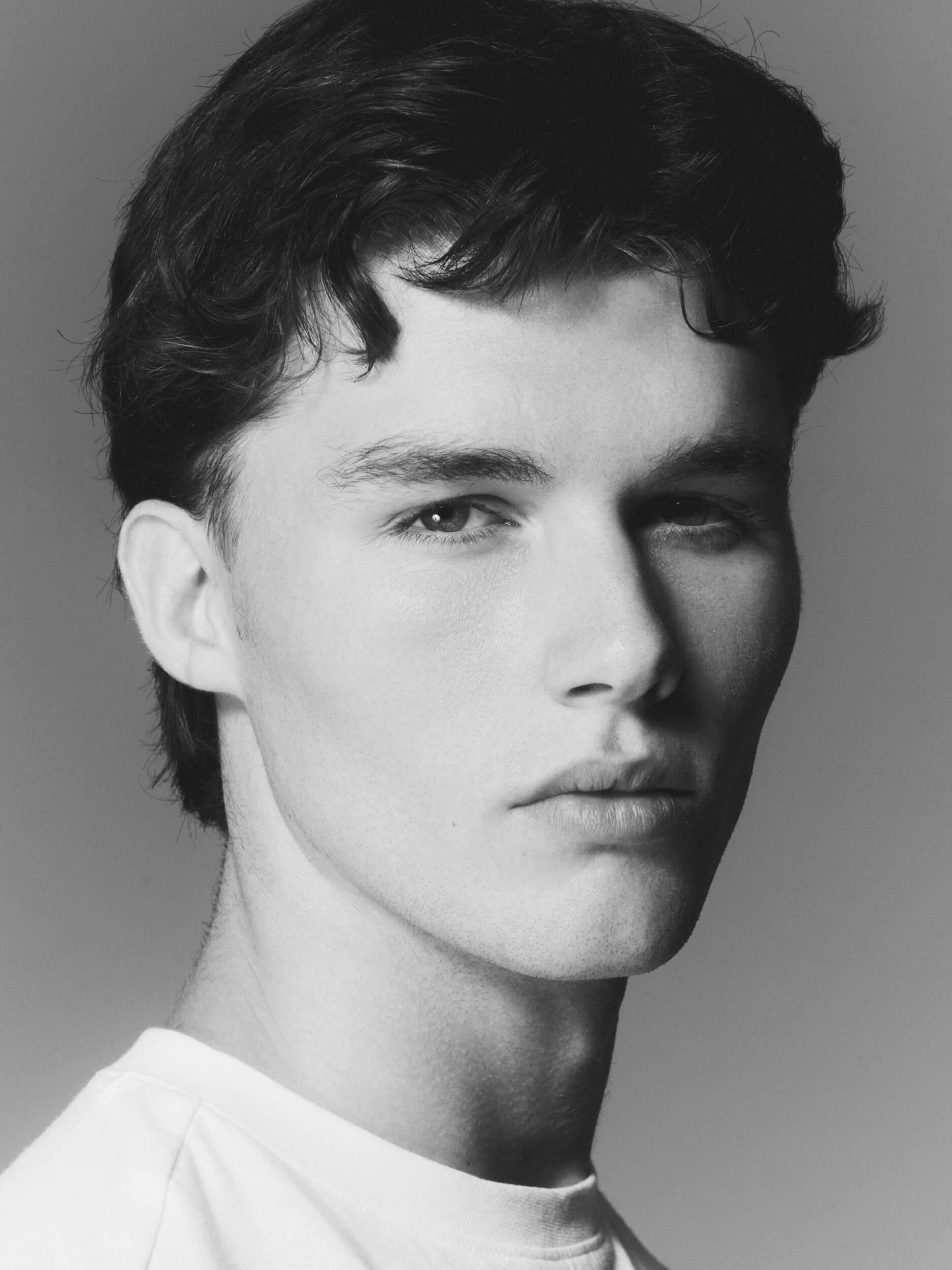 Black and white portrait of a young person with short dark hair, looking slightly to the side with a neutral expression. They are wearing a light-colored shirt and the background is plain and unobtrusive.