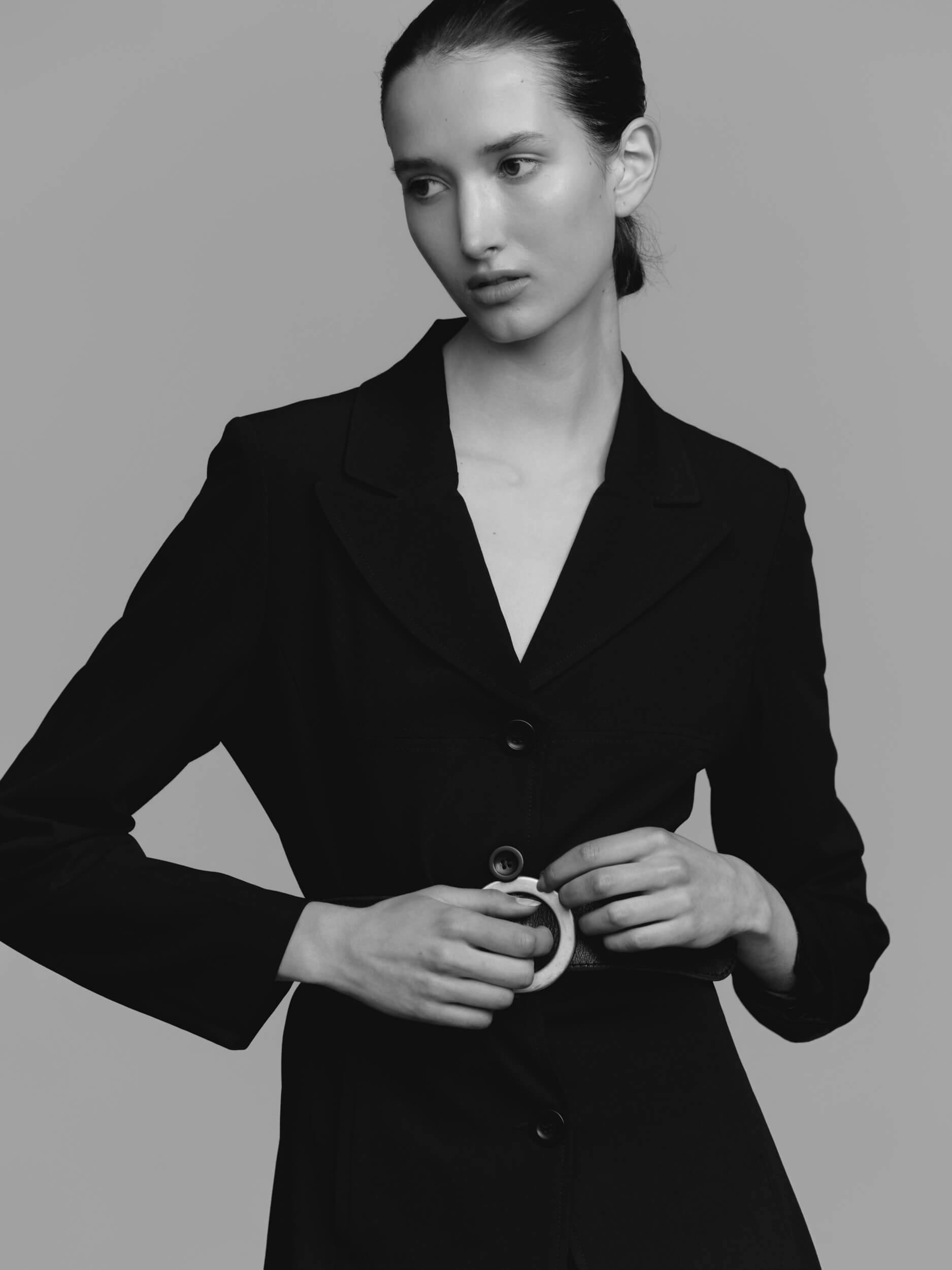 A person with long dark hair pulled back is wearing a dark blazer, standing against a plain background and looking to the side while fastening a circular belt buckle at their waist.