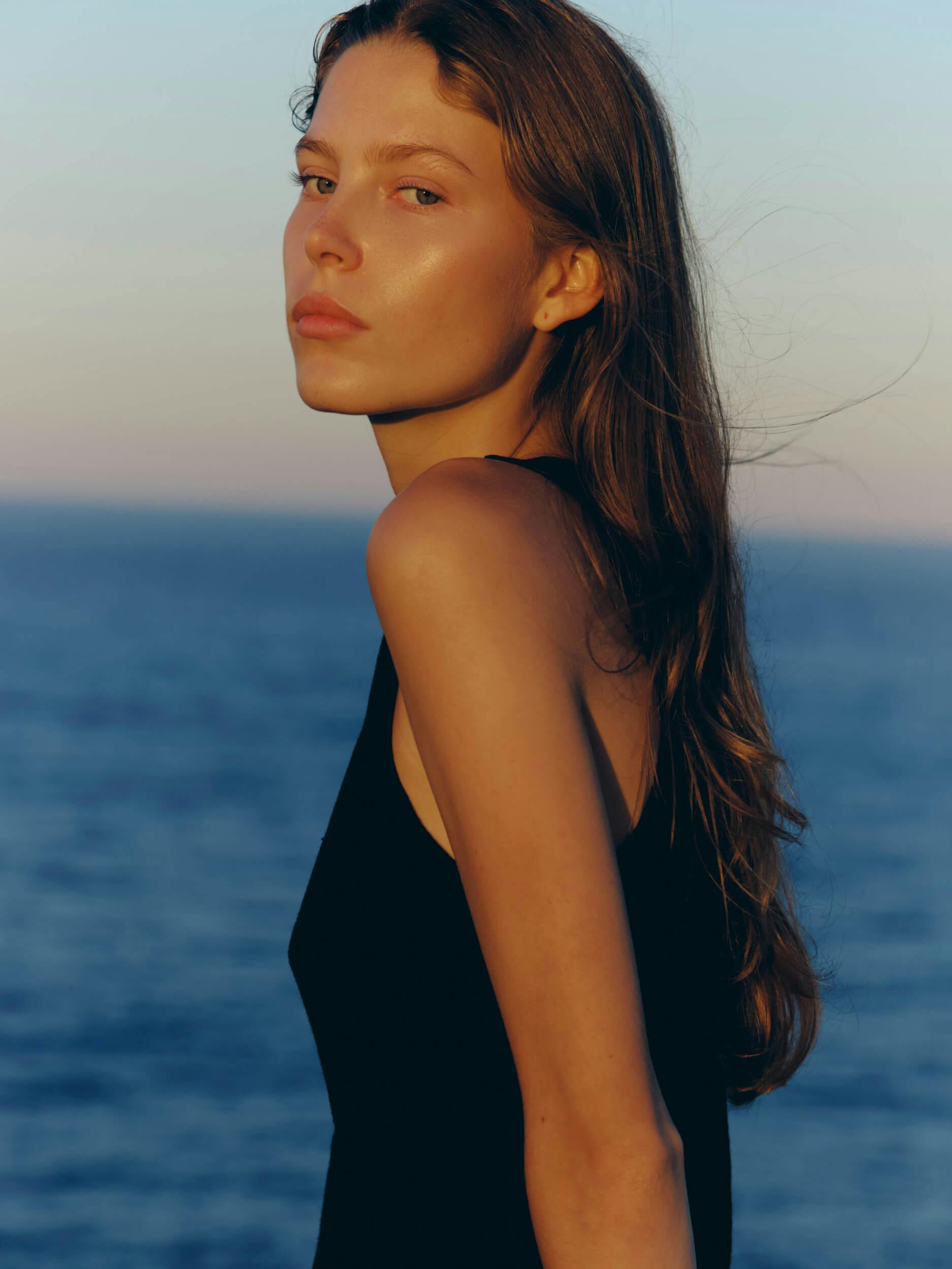 A woman with long brown hair wearing a sleeveless black top stands in profile near the ocean, with calm blue water and a pastel sky in the background. She looks toward the camera with a neutral expression.