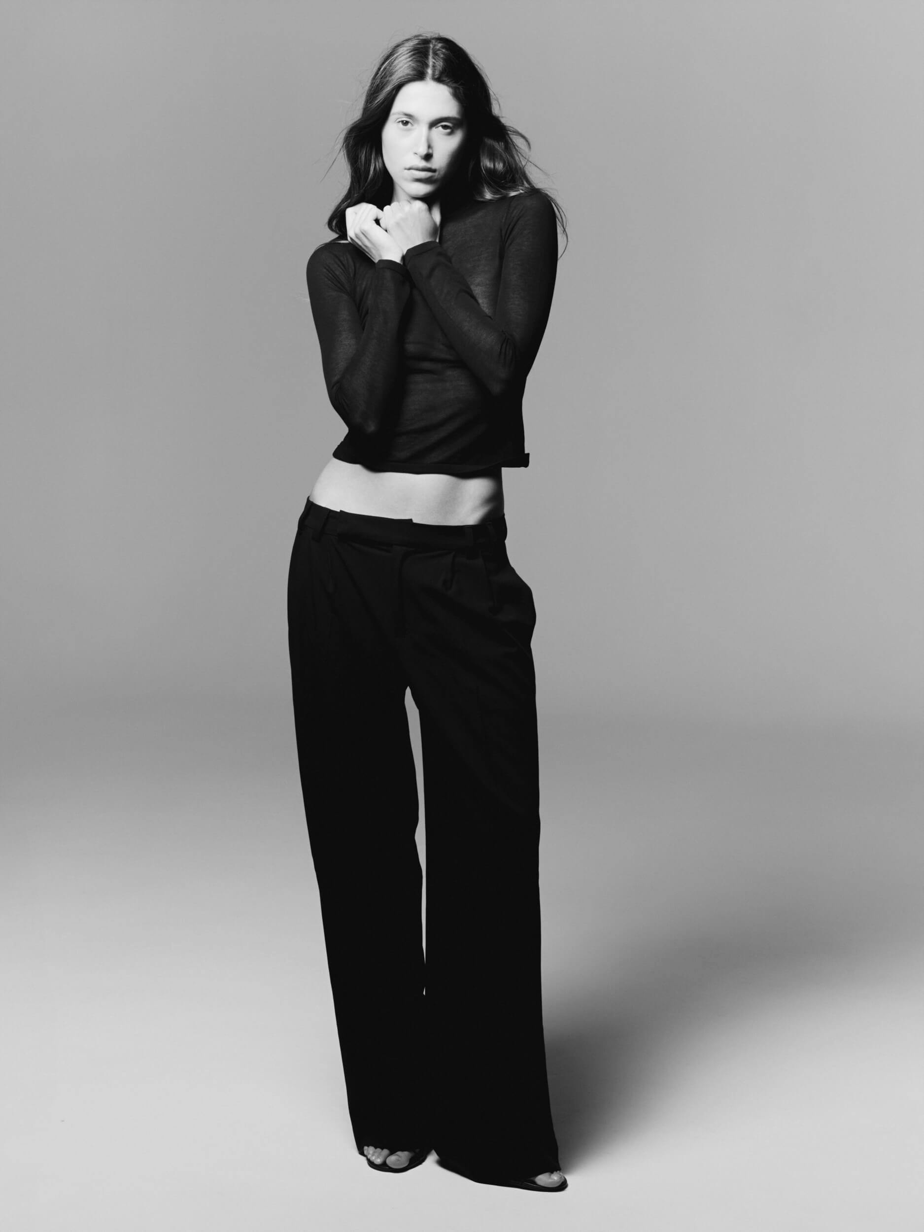 A woman stands against a plain backdrop, wearing a long-sleeve, semi-sheer black top and loose black pants. She poses with her hands near her neck, looking directly at the camera with a neutral expression.