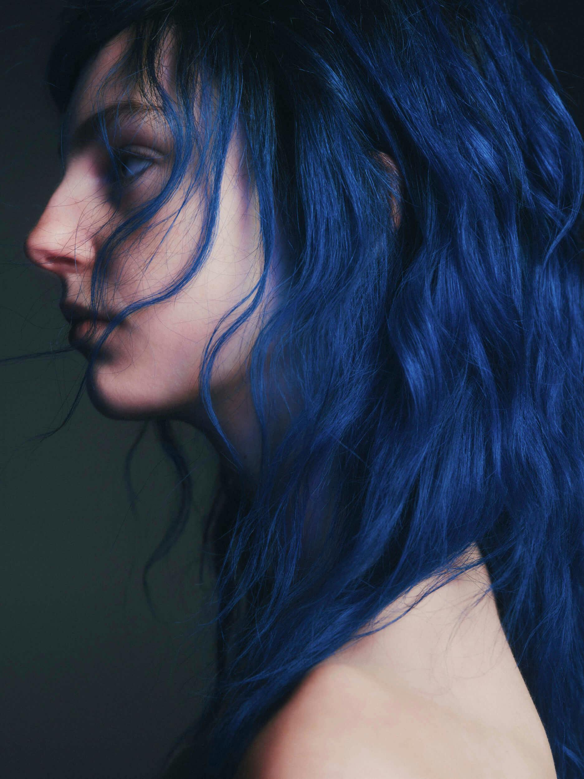 A person with long, wavy, dark blue hair is shown in a close-up side profile against a dark background. Their face is partially obscured by their hair, and their shoulder is visible.