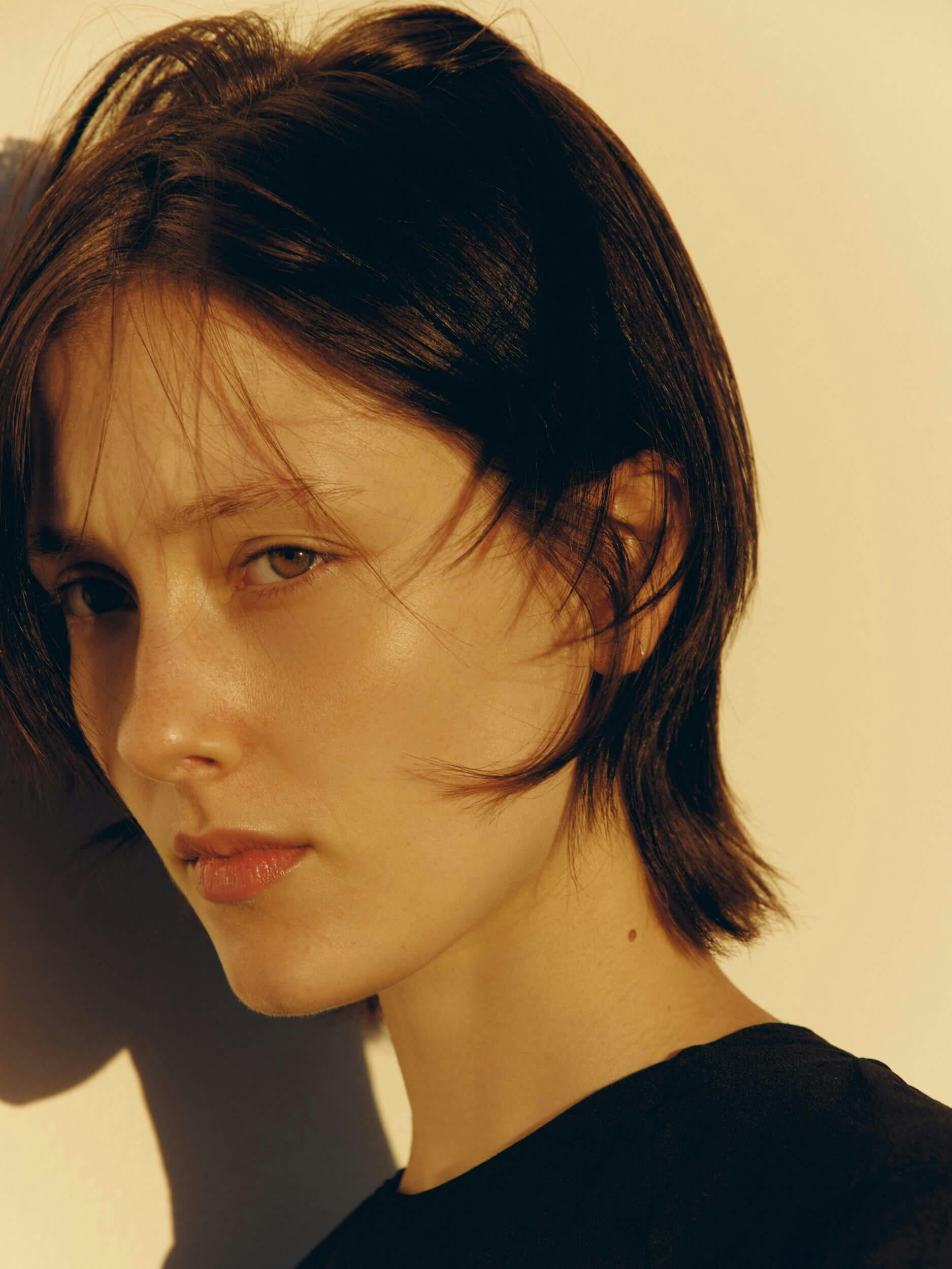 A person with short brown hair and fair skin looks toward the camera with a neutral expression. Warm light casts shadows on their face and a pale background. They are wearing a black shirt.