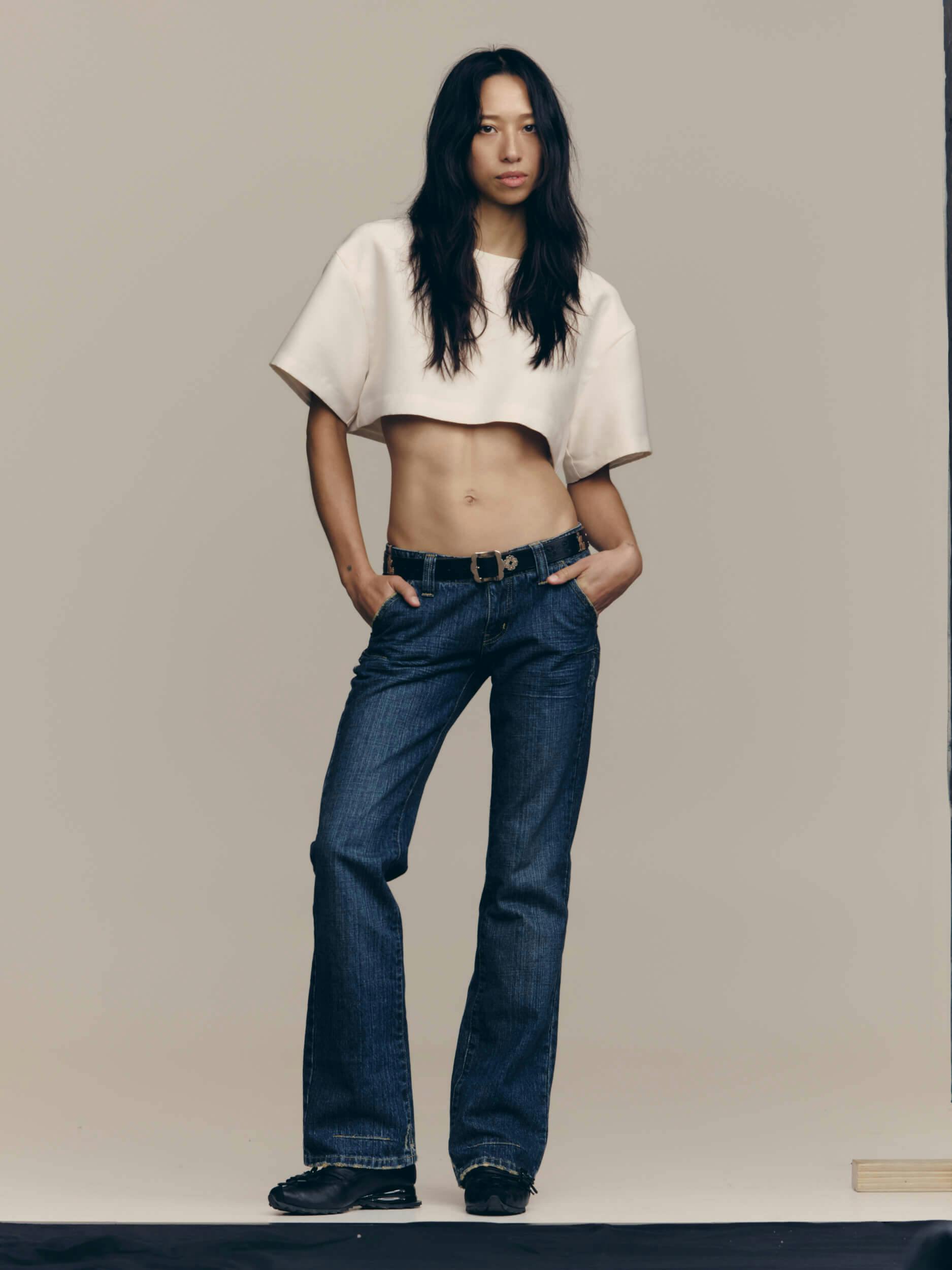 A person with long dark hair stands against a plain background, wearing a white cropped T-shirt, blue jeans, and black shoes. They have their hands in their pockets and face forward.