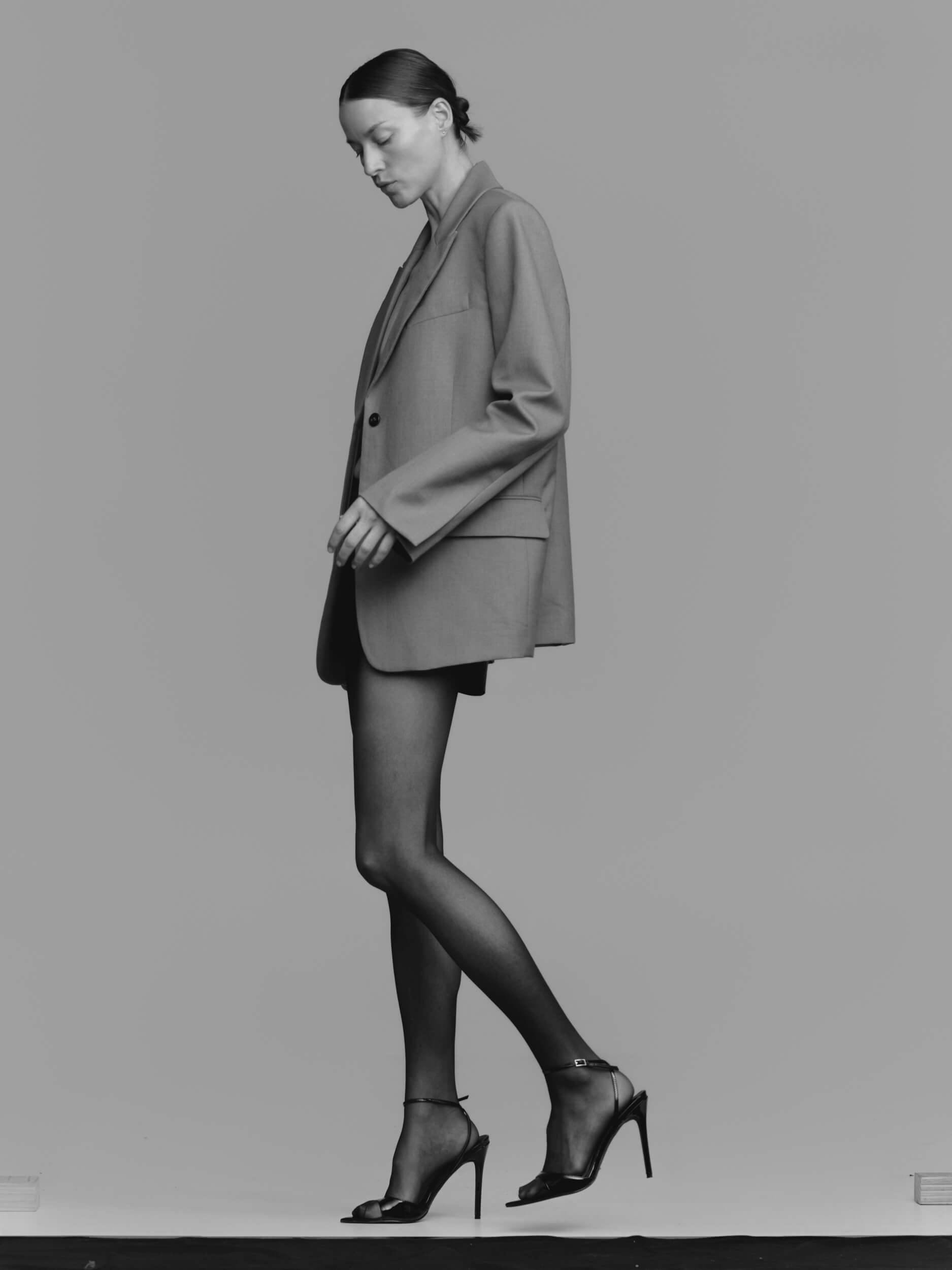 A person wearing a long blazer, sheer black tights, and high-heeled sandals stands in profile against a plain, light gray background.