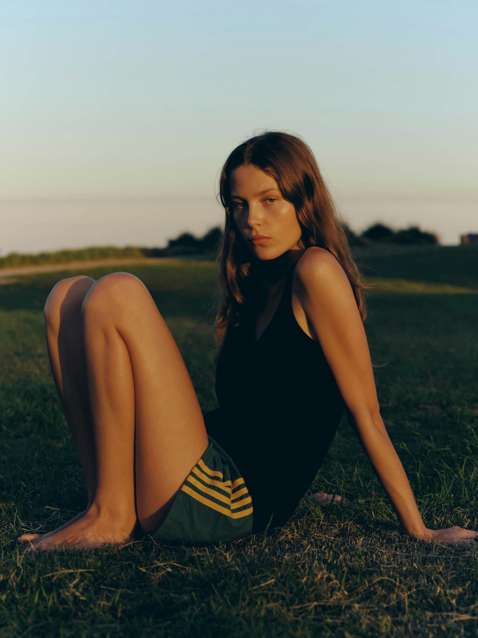 A person with long brown hair wearing a black tank top and green shorts sits barefoot on grass outdoors, with arms extended back and legs bent, in soft natural light. Trees and sky are visible in the background.