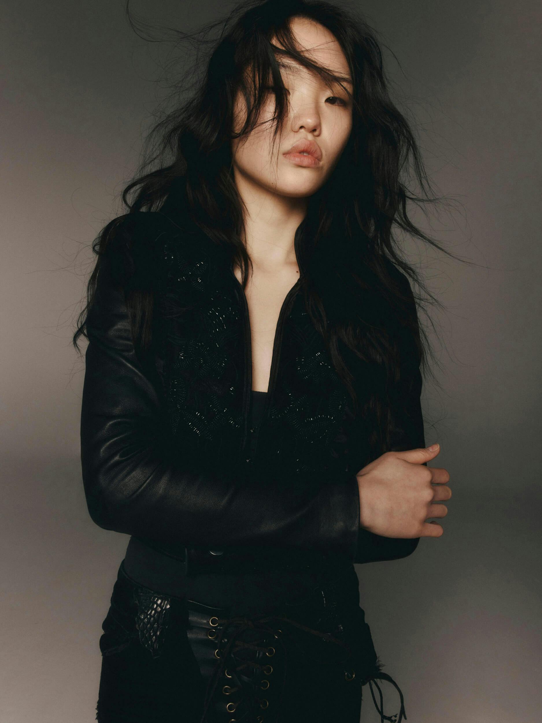 A person with long dark hair wearing a black jacket stands with arms crossed against a plain background. Their hair is tousled, and they have a neutral facial expression.