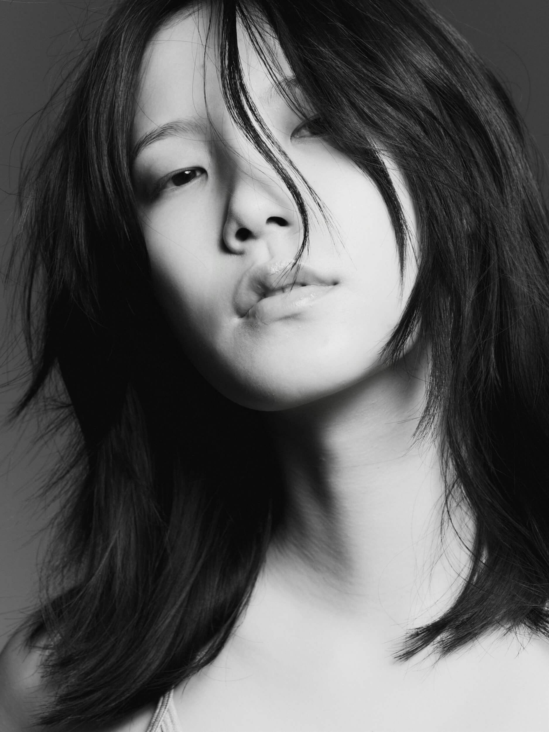 Black and white close-up photo of a young person with shoulder-length dark hair, looking at the camera with a neutral expression; some hair falls across their face. The background is plain and dark.