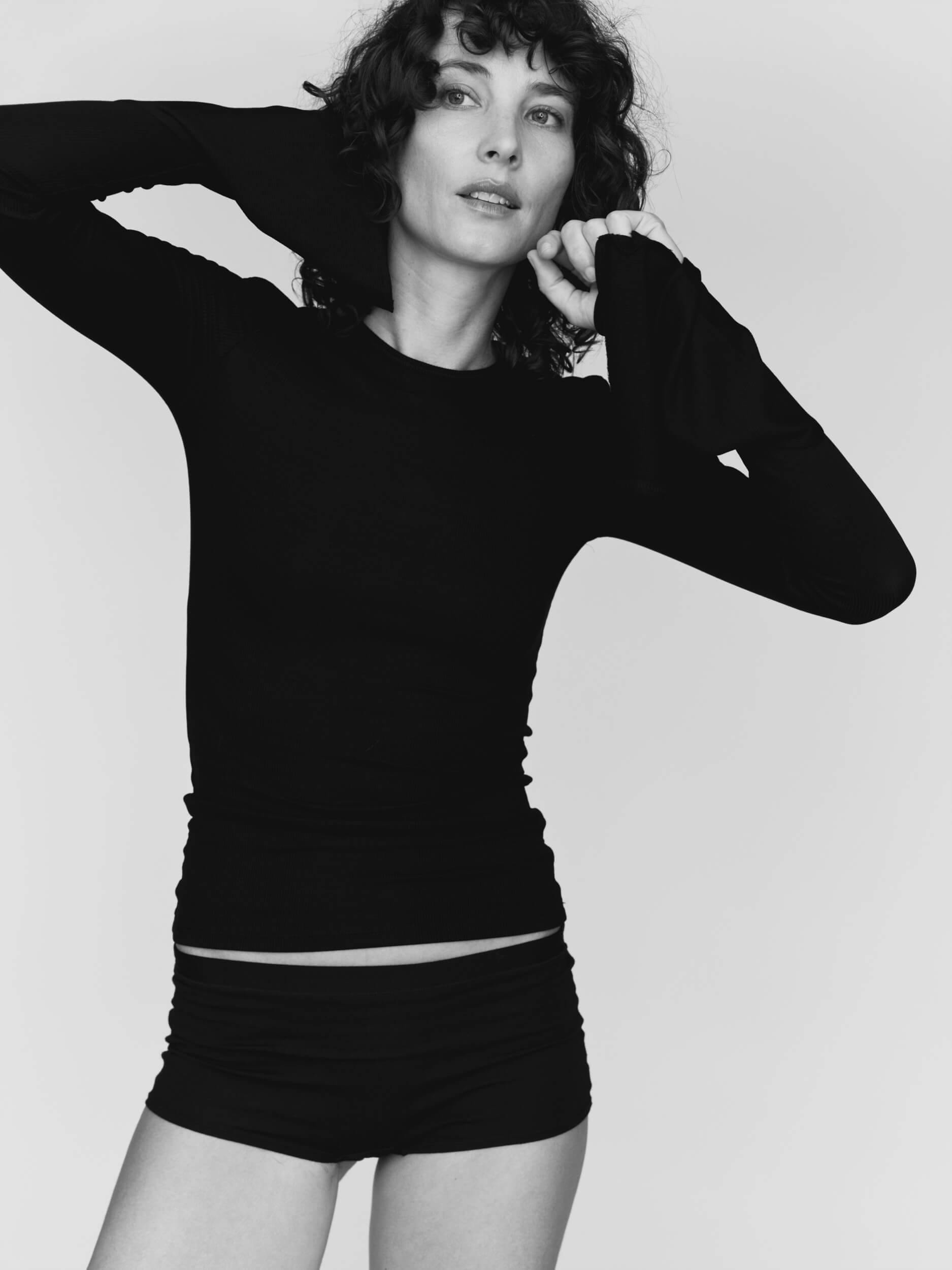 A person with curly hair wearing a long-sleeved black top and black shorts poses against a plain white background, with one arm raised and the other bent near the face.