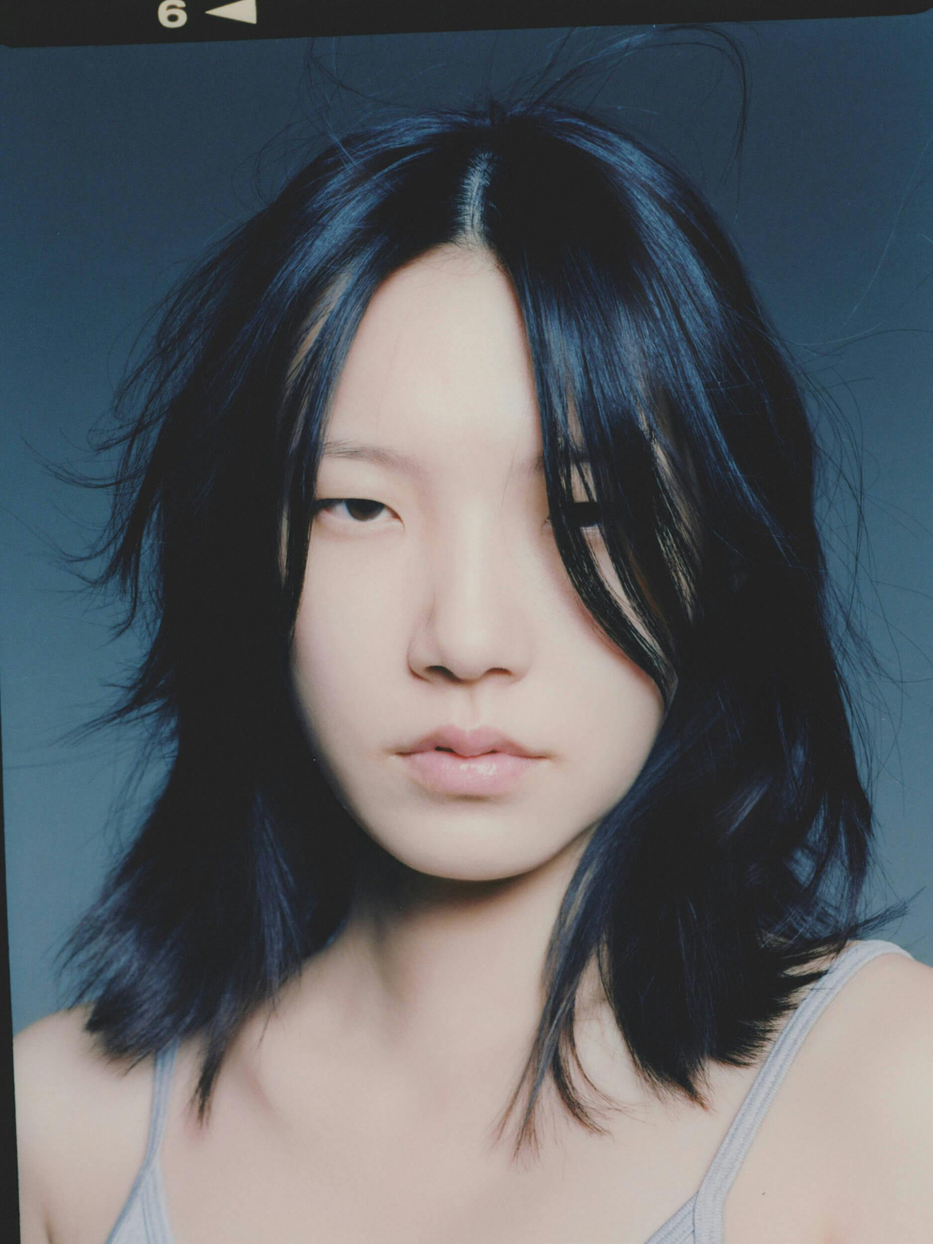 A person with medium-length, layered black hair looks directly at the camera with a neutral expression, wearing a gray tank top against a plain, blue-gray background.