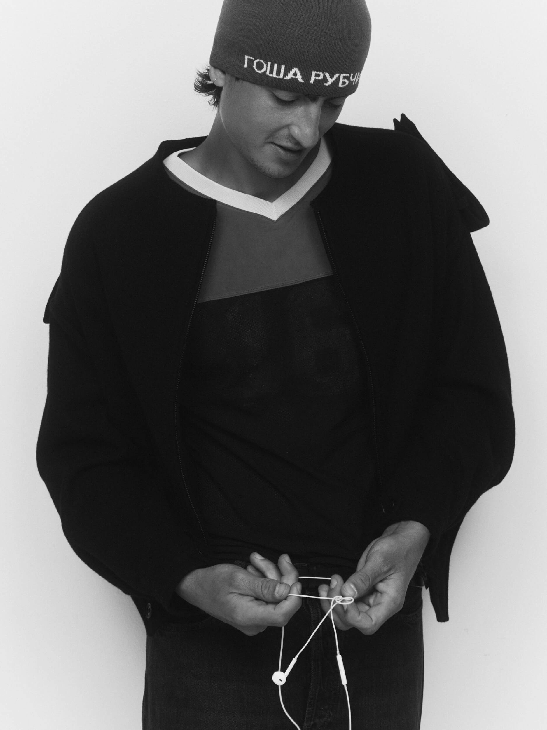 A person wearing a beanie with Cyrillic text, a dark jacket, and a t-shirt looks down while holding a pair of wired earphones against a plain background.