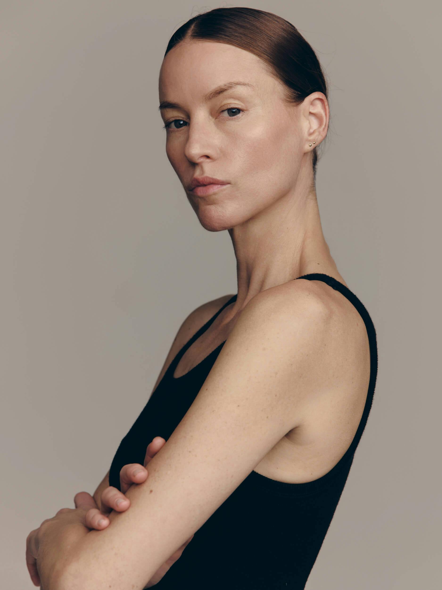 A person with fair skin and brown hair pulled back wears a sleeveless black top, standing with arms crossed and a neutral expression against a plain, light background.
