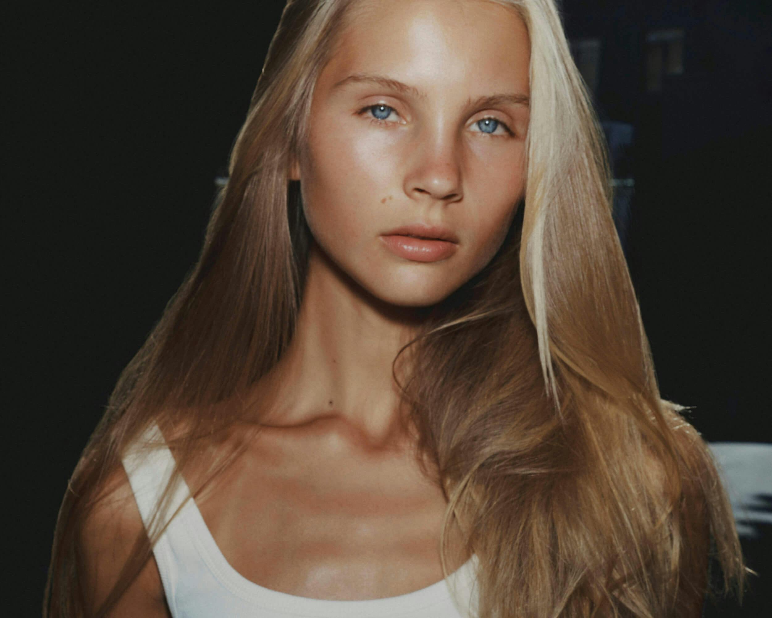 A young woman with long, straight blonde hair and blue eyes looks directly at the camera. She is wearing a white tank top and stands against a dark background.